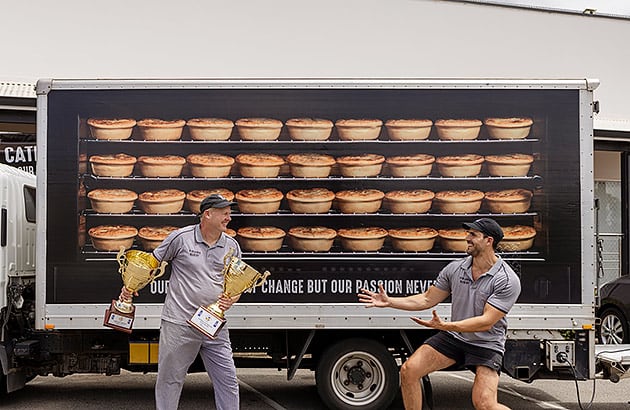 Two men standing next to a bakery truck, with one holding a trophy