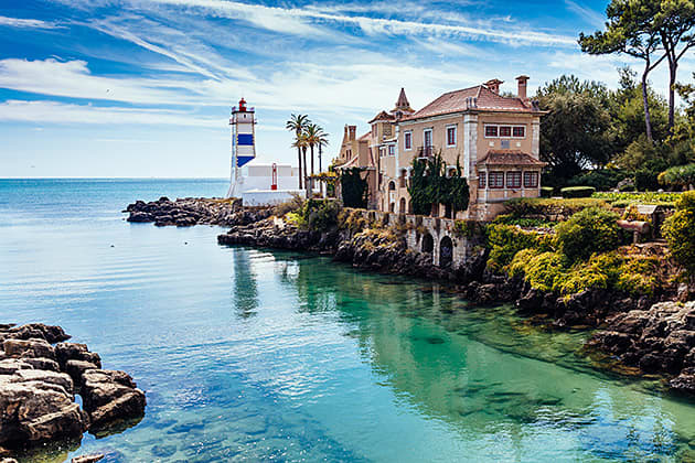The white and blue Santa Marta Lighthouse is perched on a rocky bank next to the Museum Cascais. There is crystal clear green water running close to these buildings, with rocky outcrops and green foliage surrounding.