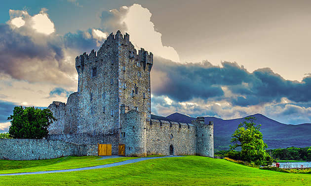 A grey stone castle is surrounded by bright green lawn, with mountains and dark grey clouds behind in the distance.
