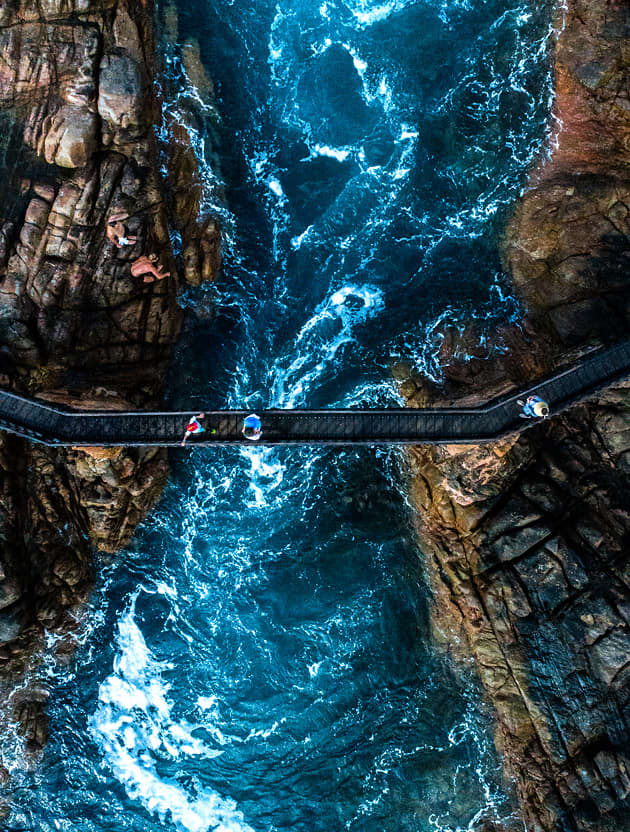 An arial view of three people walking along a narrow bridge while the ocean swirls below them at Canal Rocks in Yallingup.