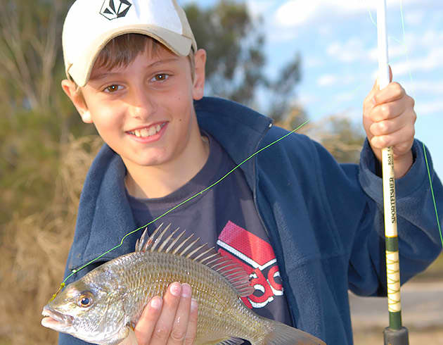 A young boy with a fishing rod holds up his catch