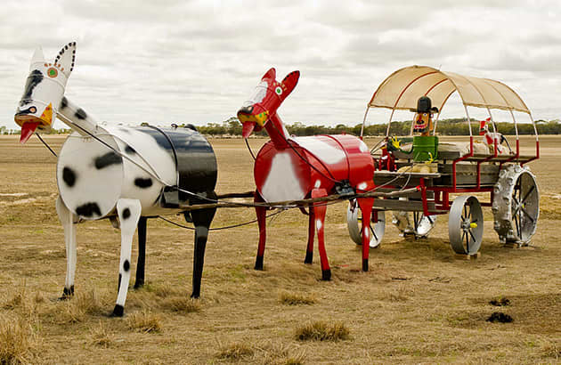Two tin horses are positioned in front of a tin man in a tin wagon in the middle of a field.