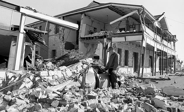 A man is touching the outside of a crumpled car, which is buried under a pile of bricks from a neighbouring building that has been destroyed in the Meckering earthquake