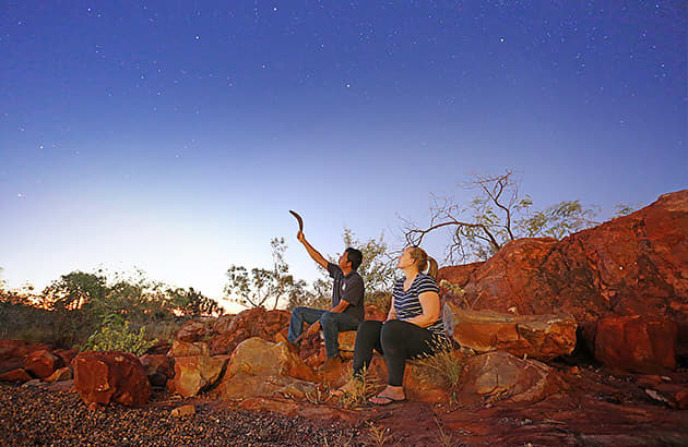 A traditional owner holding a boomerang is sitting next to a woman and pointing at the night sky