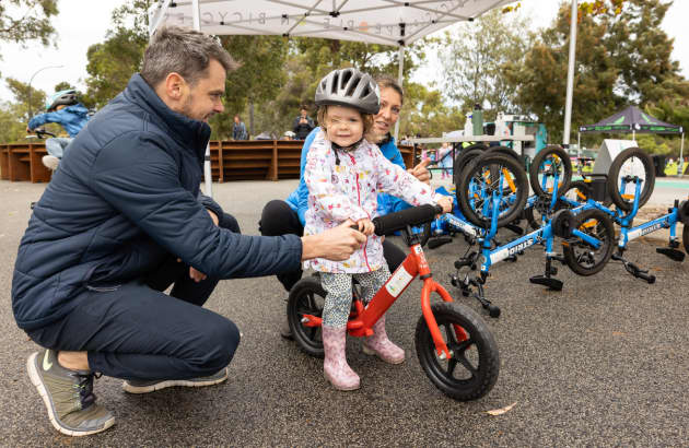 A young child sitting on a childrens' bike with parents crouching either side