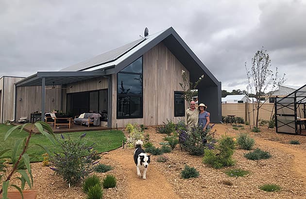 A couple standing outside their home with a dog walking in the foreground