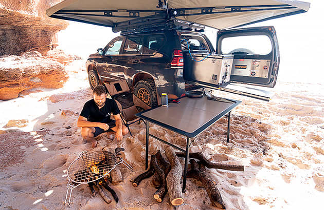A man cook tending a campfire under an awning near a 4WD