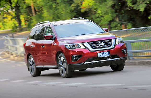 A red Nissan Pathfinder driving past lush green roadside foliage.