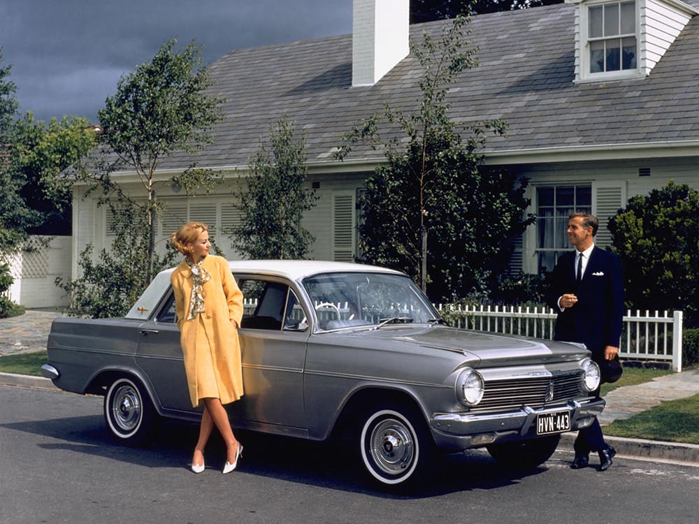 A grey 1960s Holden EH outside a family home with a male female on either side