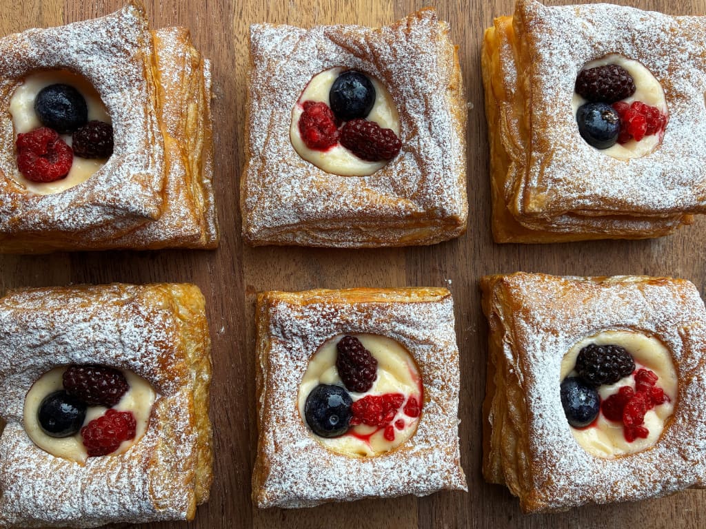 Six square, golden-brown Danish pastries are arranged in two rows of three on a wooden surface.Each pastry has a puffy, multi-layered flaky crust heavily dusted with powdered sugar around the edges. The center of each pastry features a round well filled with creamy yellow custard, topped with a small cluster of fresh berries, including blueberries, raspberries, and blackberries. The view is from directly overhead.