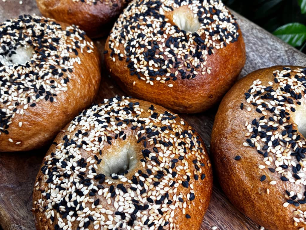 A close-up, angled shot shows several freshly baked bagels resting on a wooden board.The bagels feature a glossy, golden-brown crust and are generously topped with a mixture of white and black sesame seeds. The focus is sharpest on the bagel in the bottom center, while the surrounding bagels blur slightly into the background, showcasing their plump texture and dense structure.