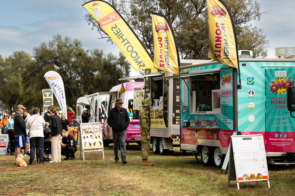 A row of food trucks parked on a grassy field at an outdoor community event with people walking by.