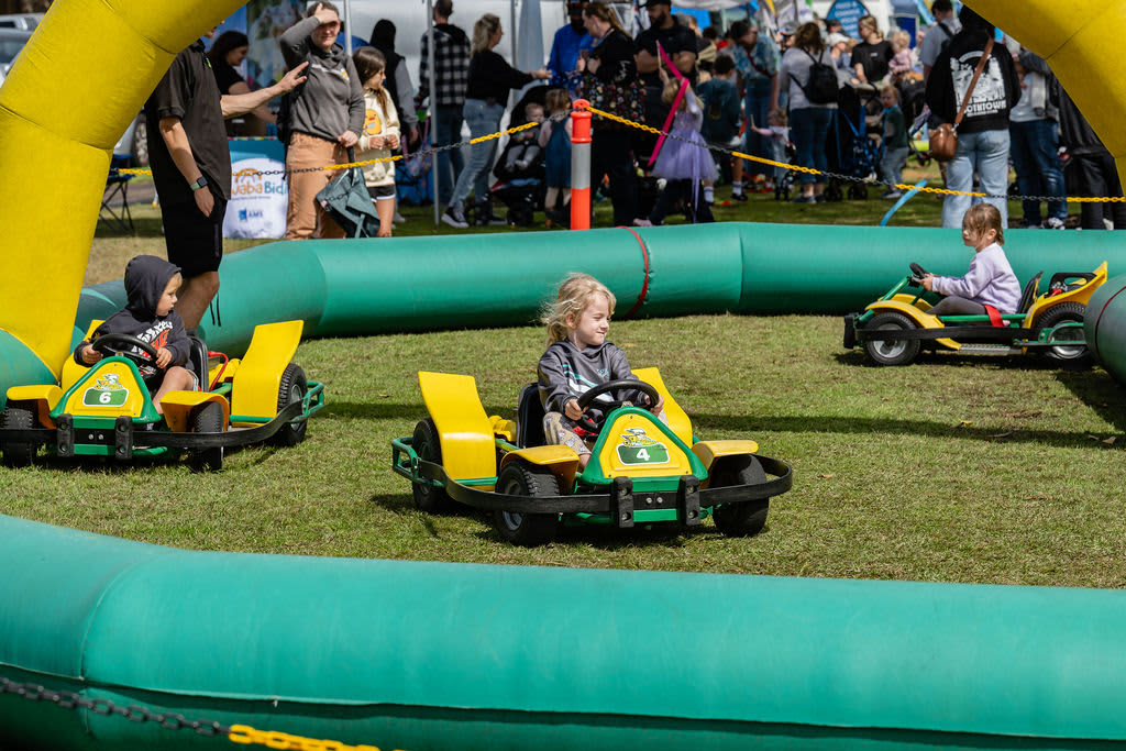 A young kid with blonde hair at a community event riding a little go kart in an inflatable race track.