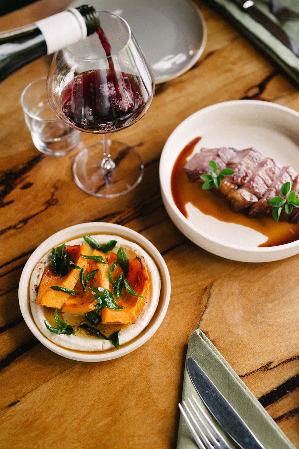 Two plates of food on a wooden table, with red wine being poured into a glass.