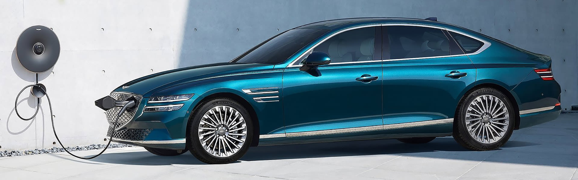 A blue Genesis G80 Electrified being charged with a white concrete wall in the background