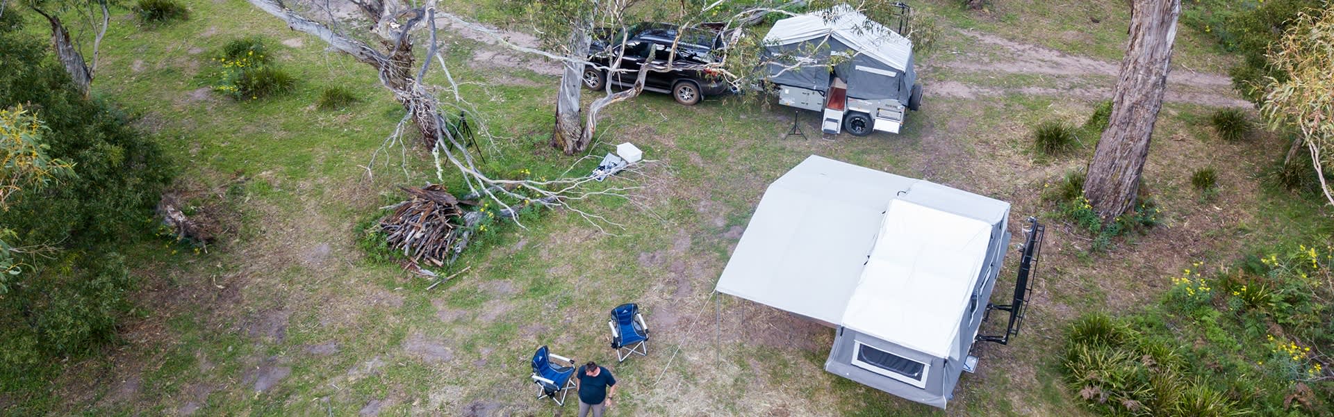 Overhead shot in a forest with two caravans, a black car and a person setting up two camping chairs.