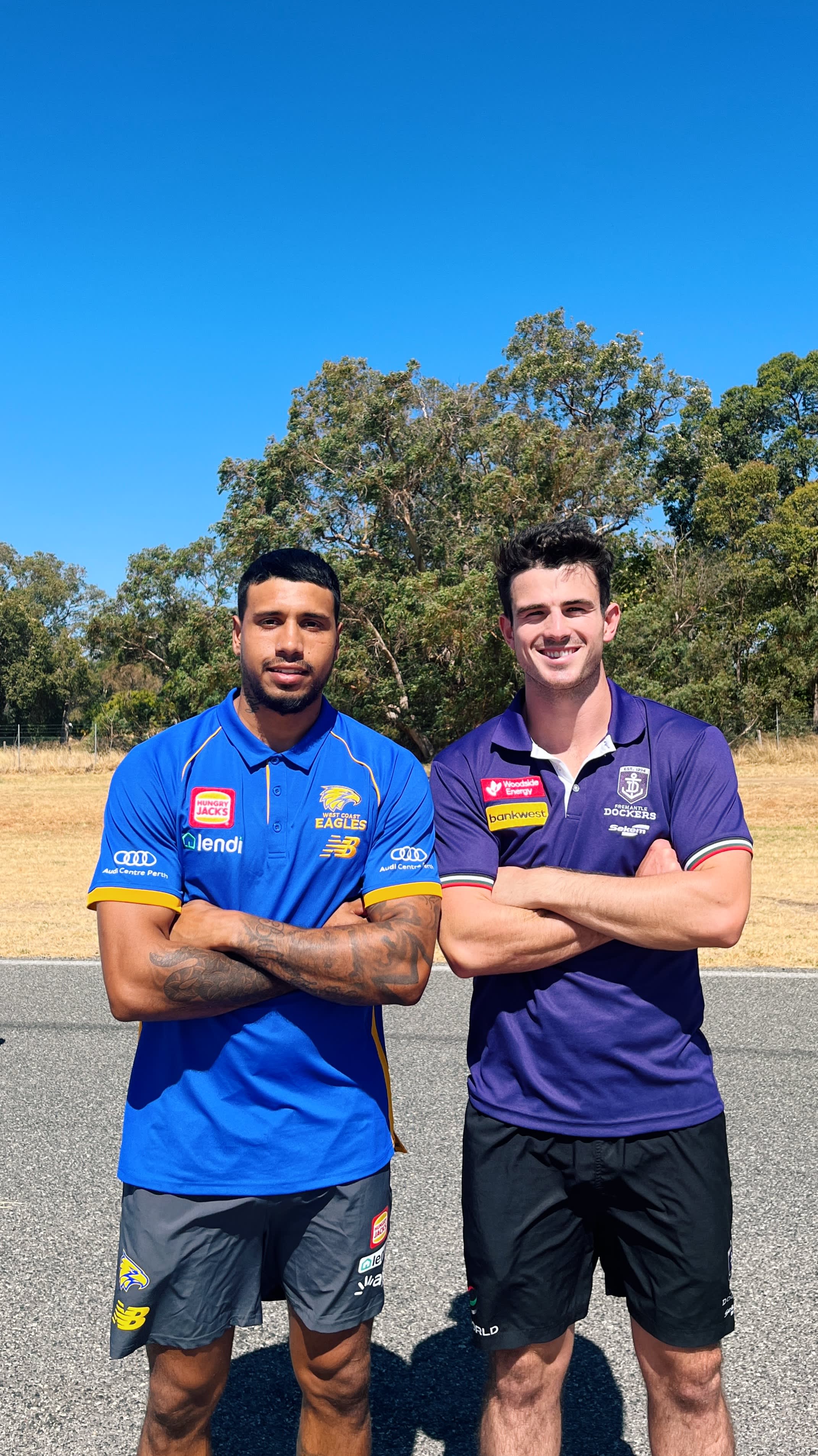 Two Aussie rules football players standing side by side in bush setting.