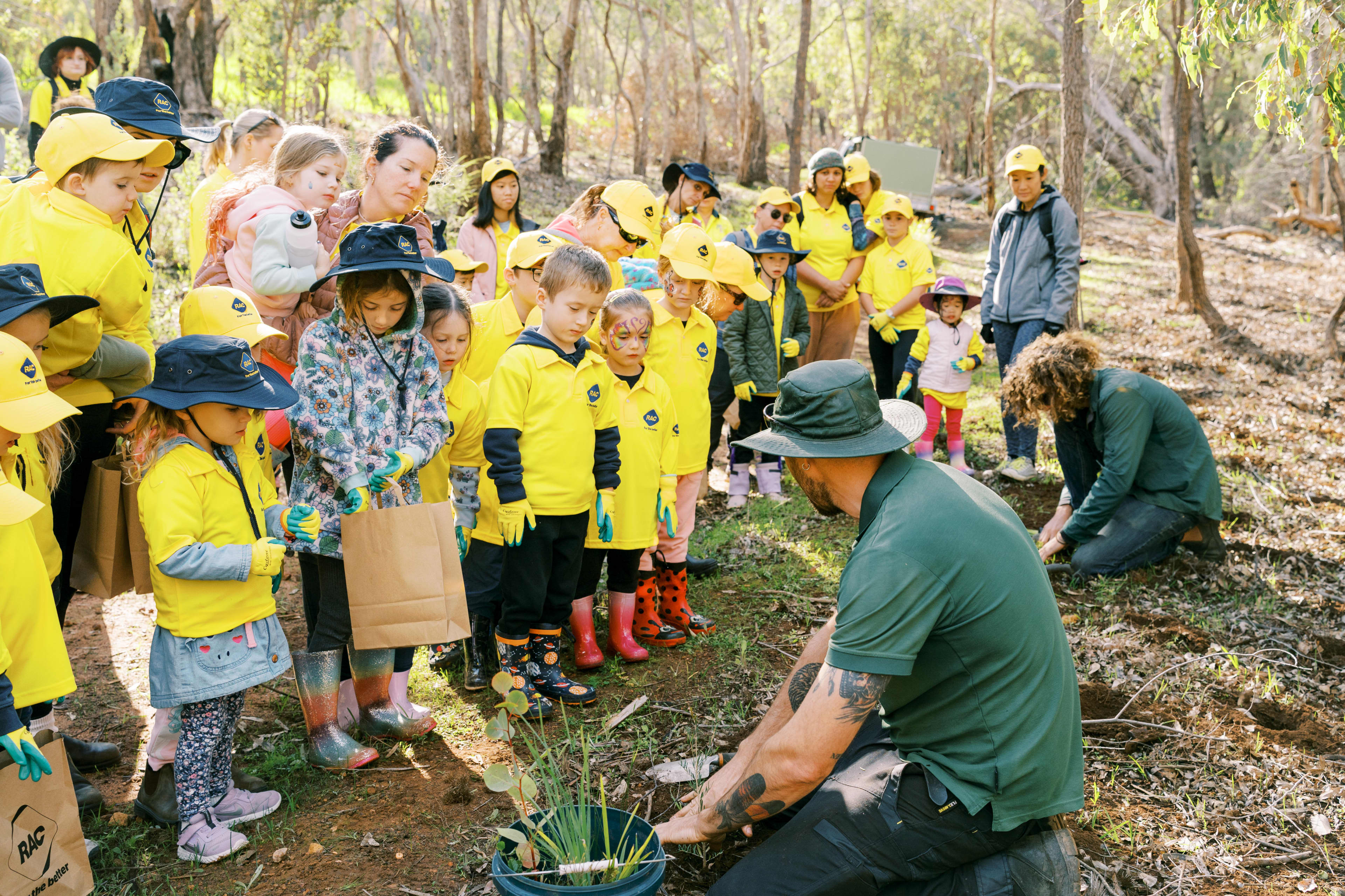 Group of primary school kids gathered around an adult showing them how to plant trees in a bush setting.