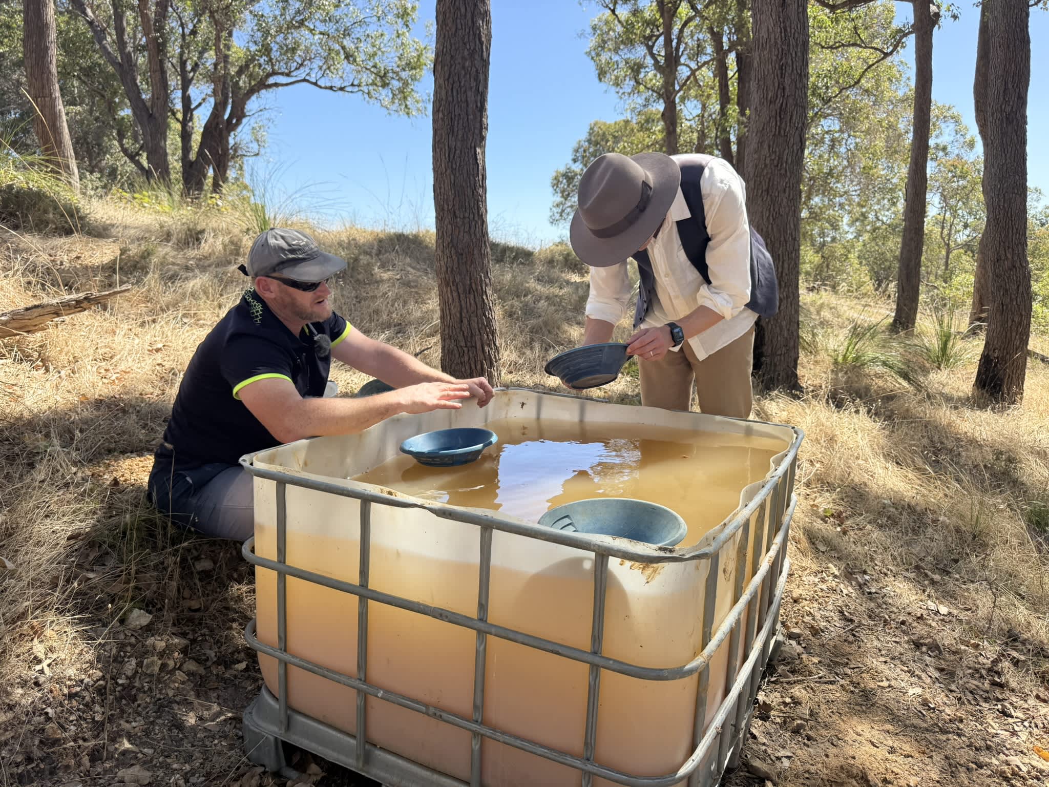 Two men are bending over a large tub filled with muddy water panning for gold