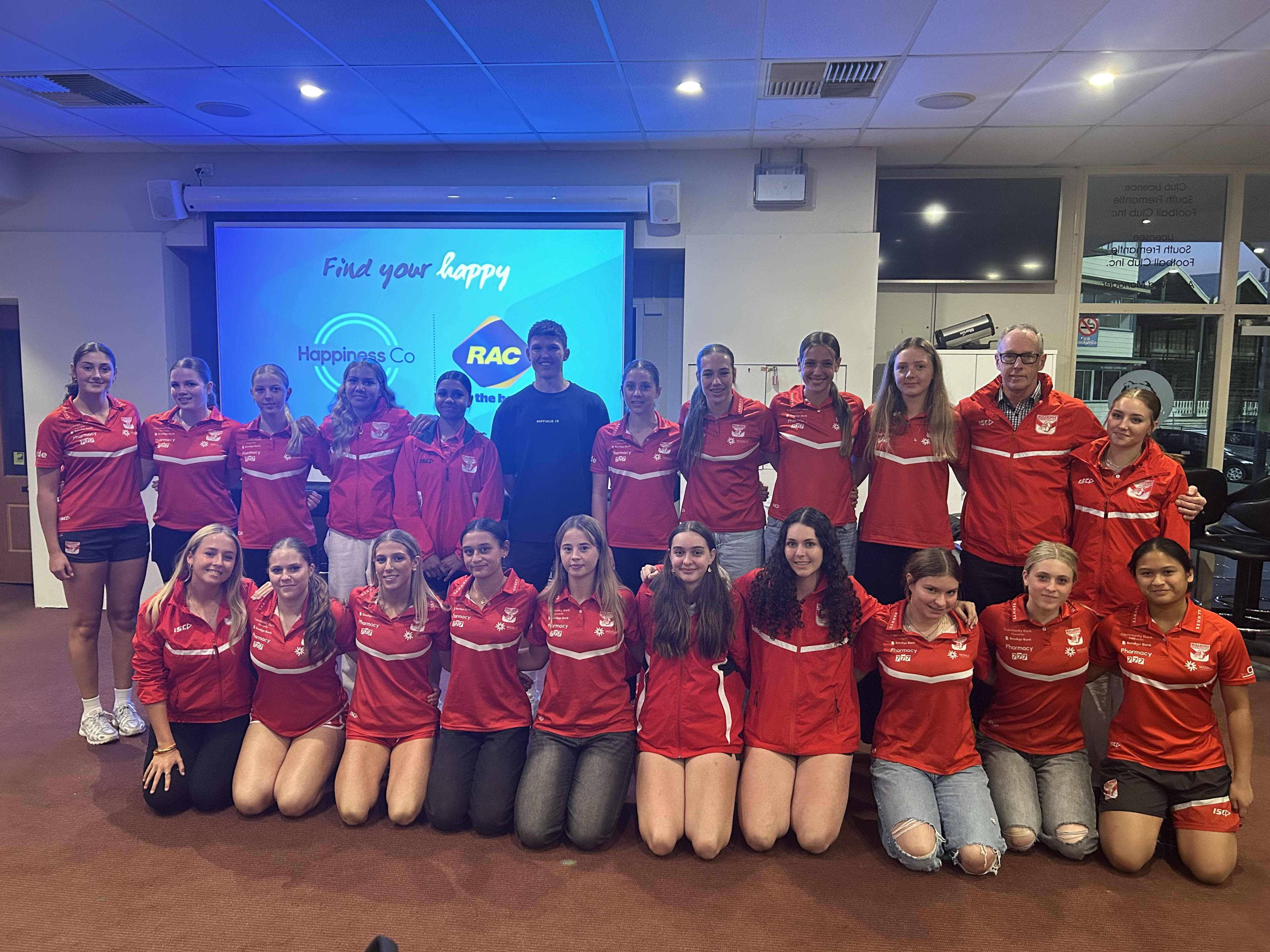 A team of women wearing red team colours posing for a photo in a seminar room