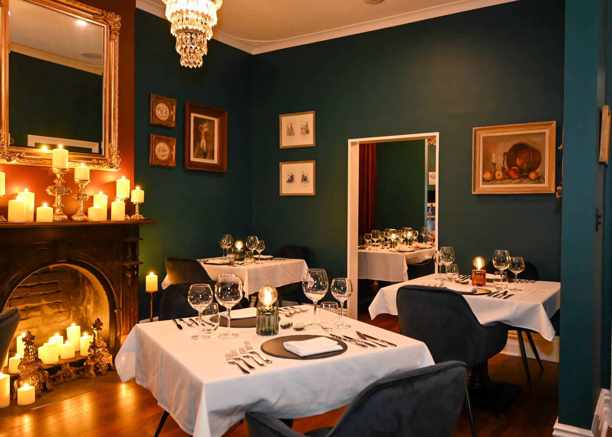A moody dining room with candles on the fireplace, a chandelier and tables set with white linen