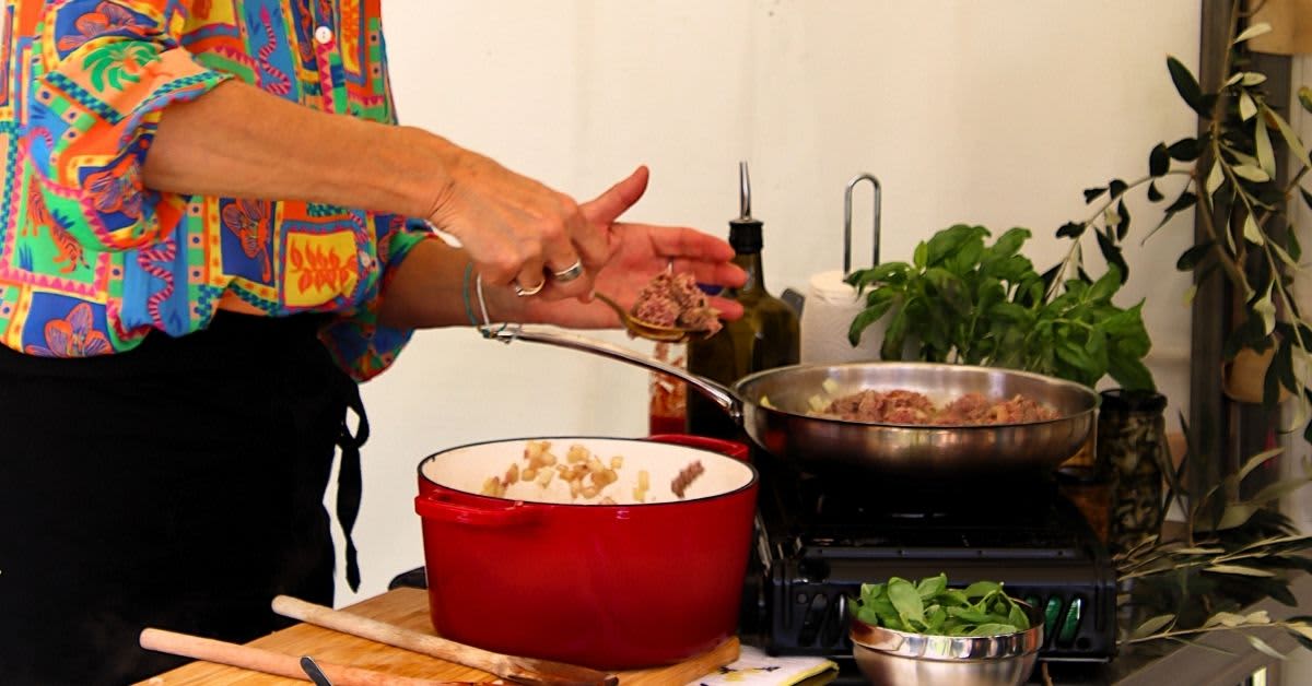 A person prepares food in different pans in live cooking demonstration with bowl of herbs and mince cooking on a pan