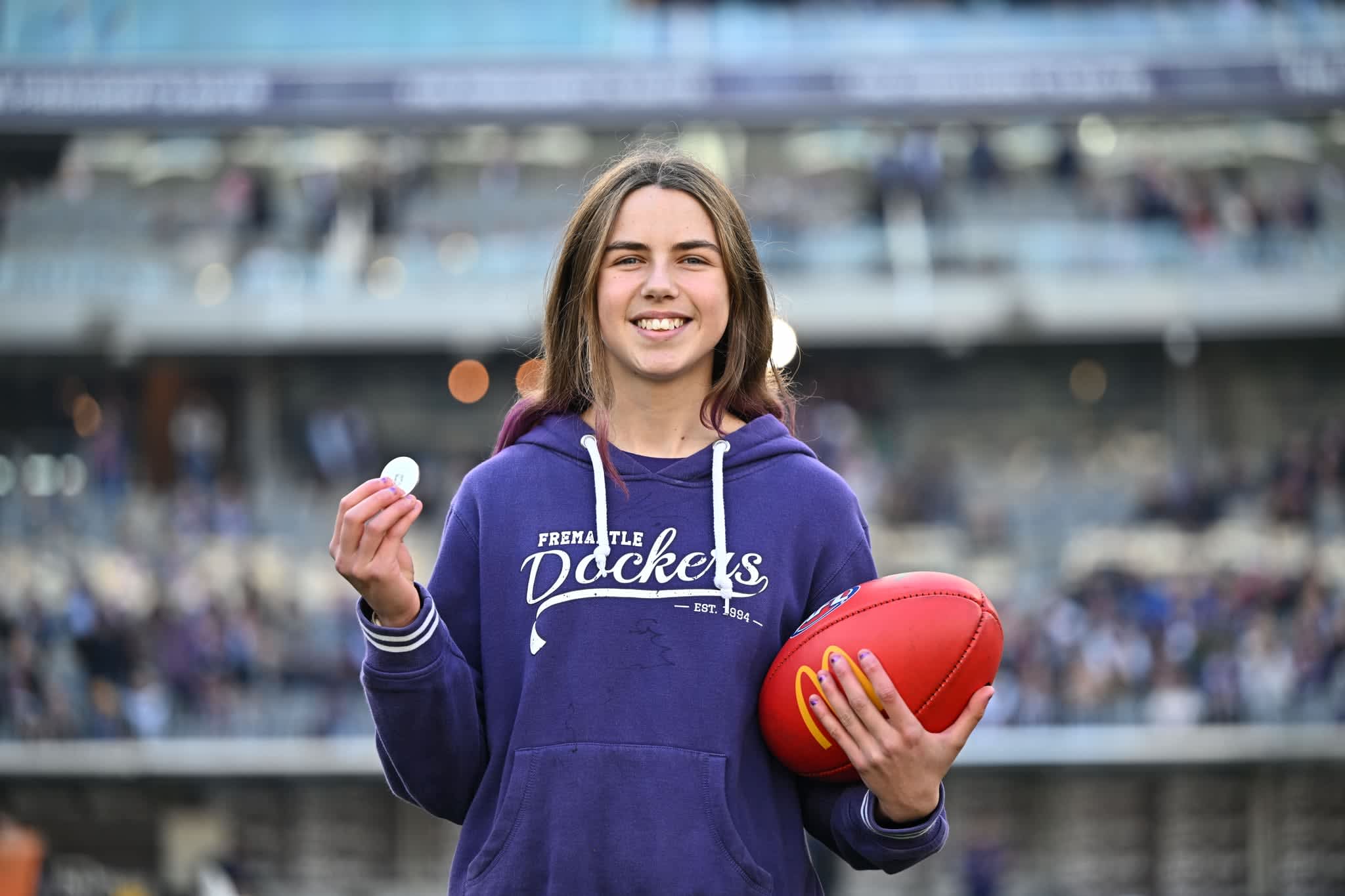 Image of the RAC Derby 61 coin toss winner, wearing a purple fremantle dockers jumper and holding the coin in one hand and the red football in the other.