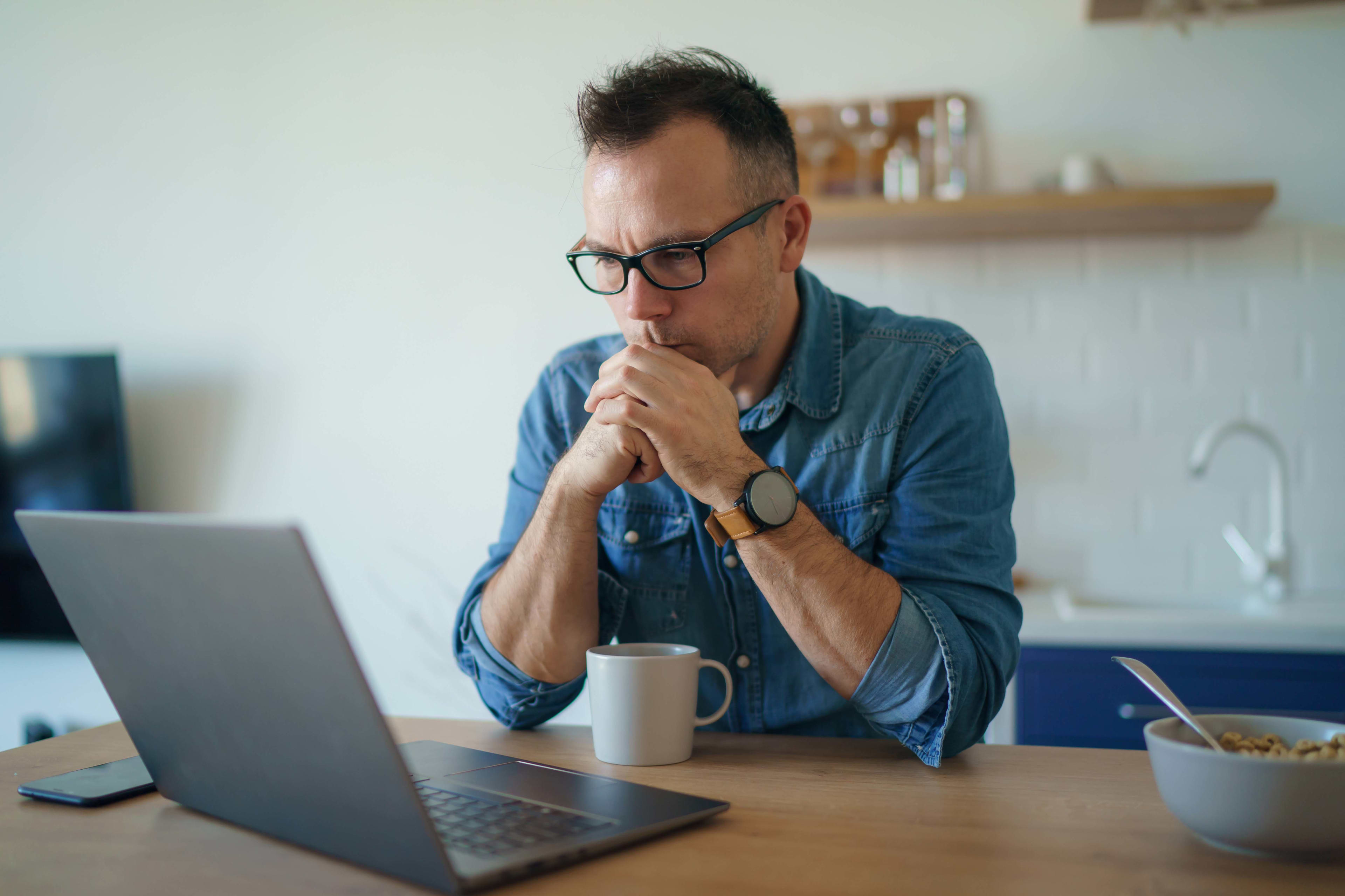 A man is sitting in his kitchen looking at a laptop screen
