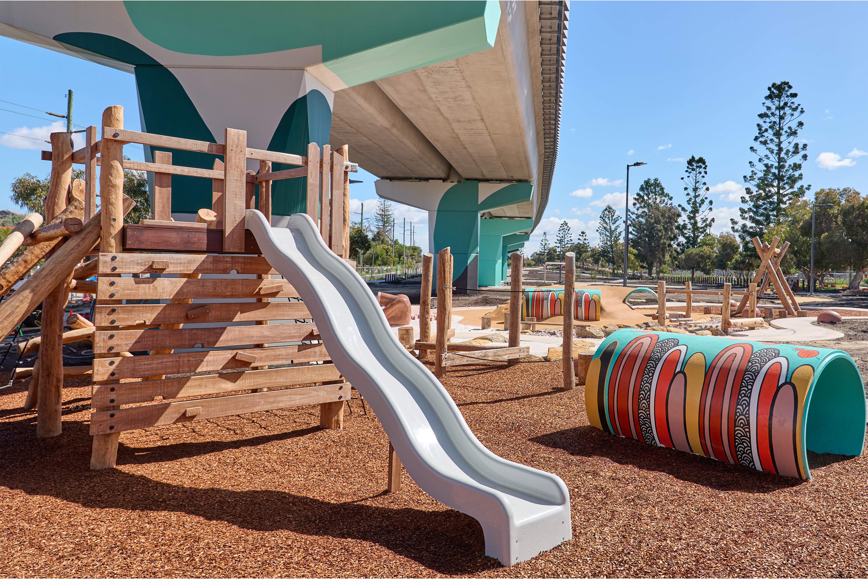 Slide underneath elevated rail line with other nature play equipment and a colourful play tunnel