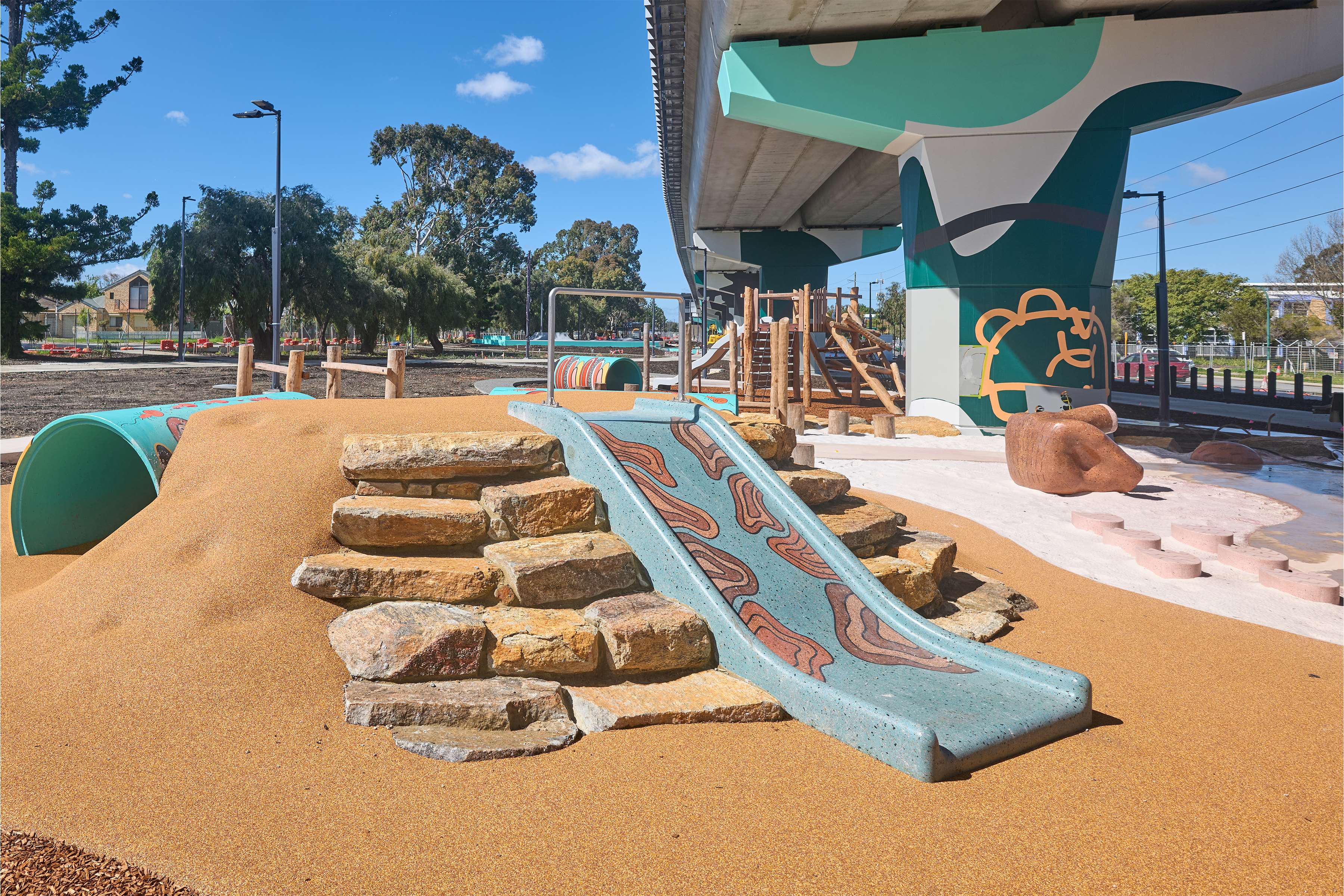 A slide surrounded by rocks and nature play equipment located underneath elevated rail line