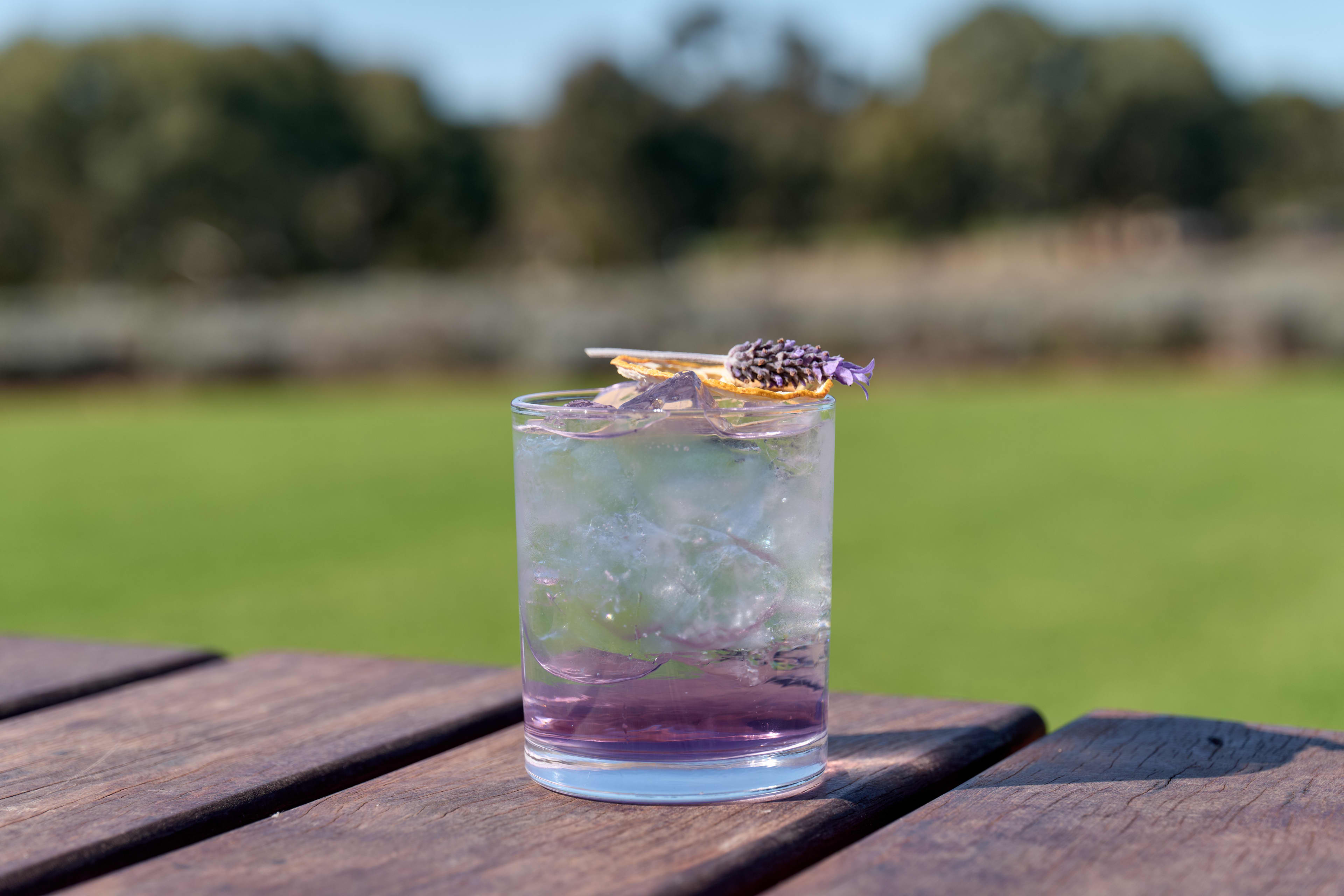 Purple-coloured glass on table with green fields in background and a sprig of lavender on top