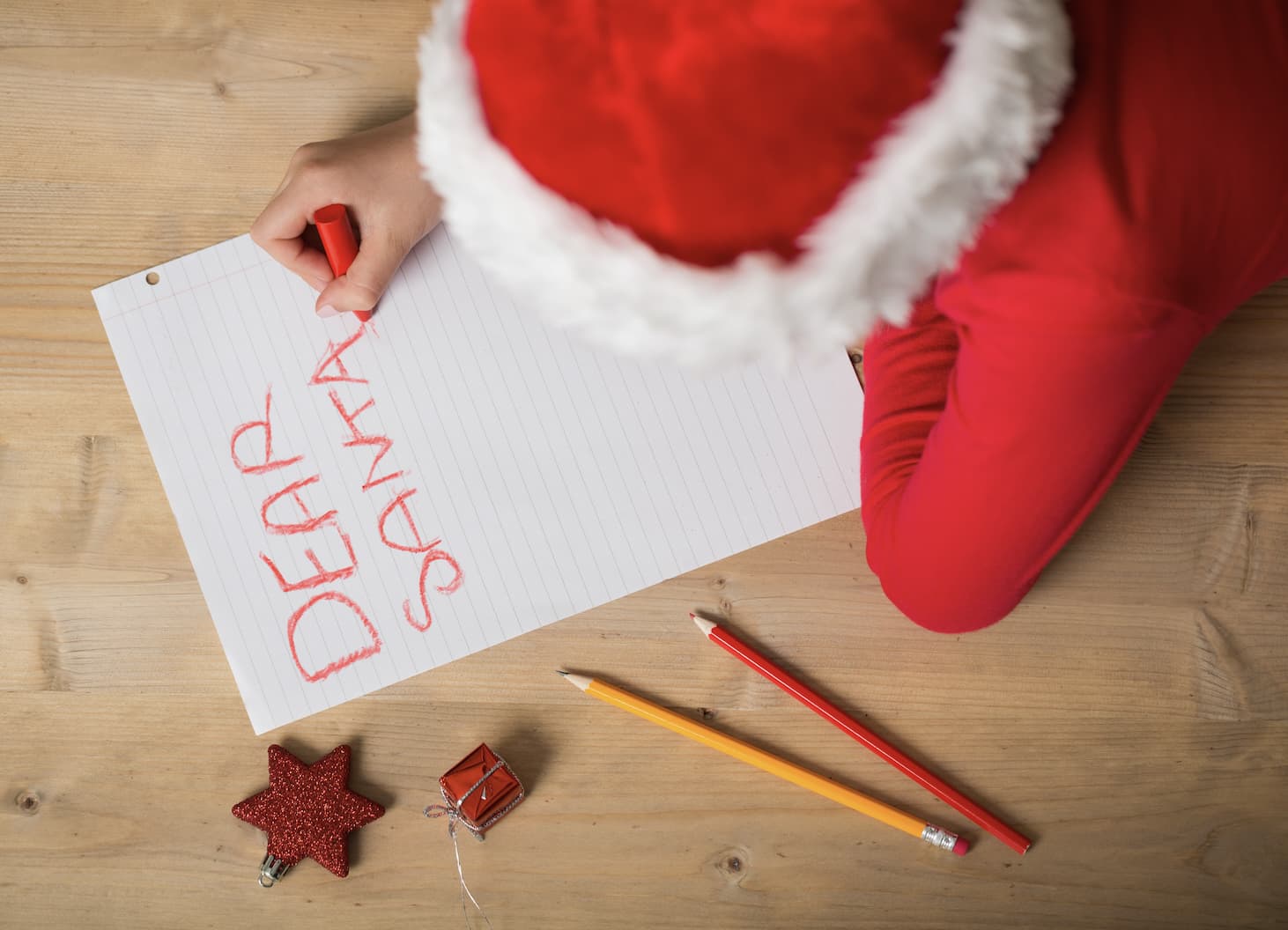 Young person lying on floor with red crayon in Santa suit writing a letter to Santa