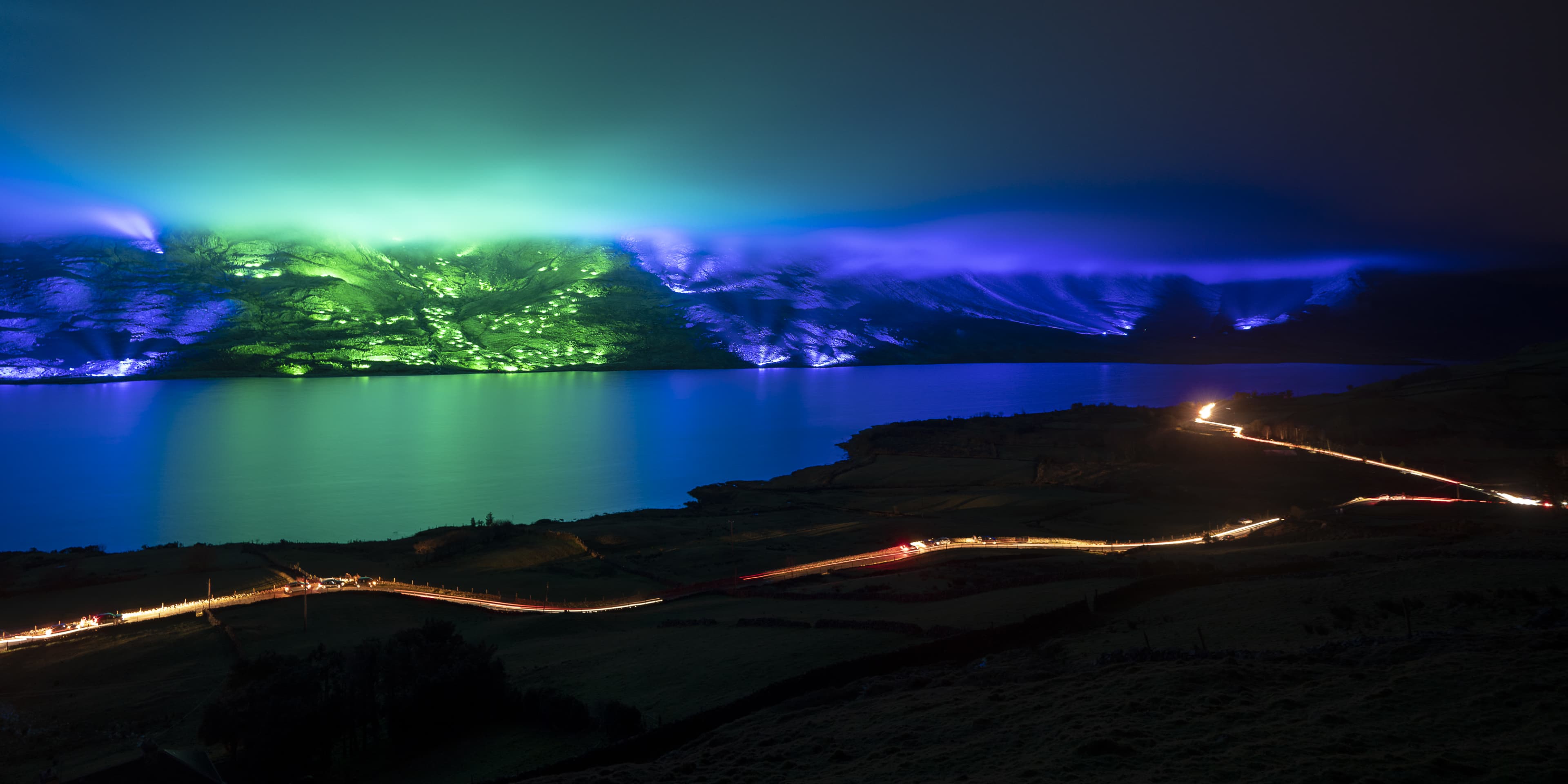 Mountains of Ireland illuminated in green and blue lights with road in foreground at night