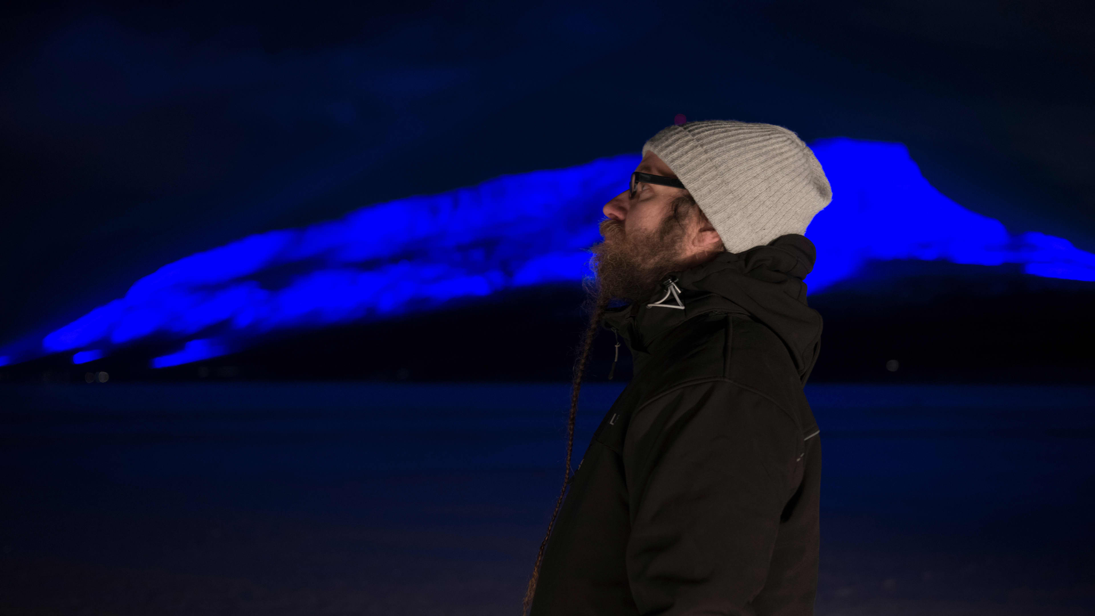 Finnish artist Kari Kola wearing a beanie and a black jacket standing side-on in front of blue illuminated mountain at night