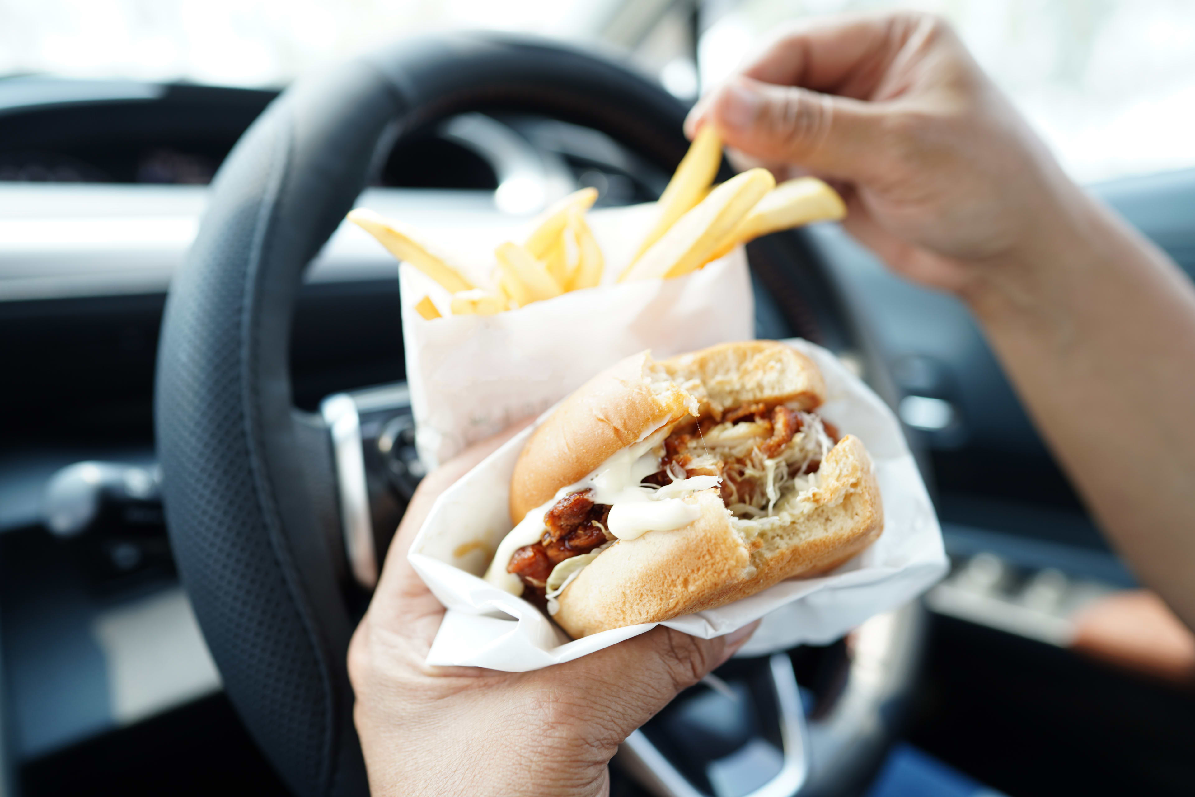 A close up of a person driving while handling a hamburger and a packet of chips