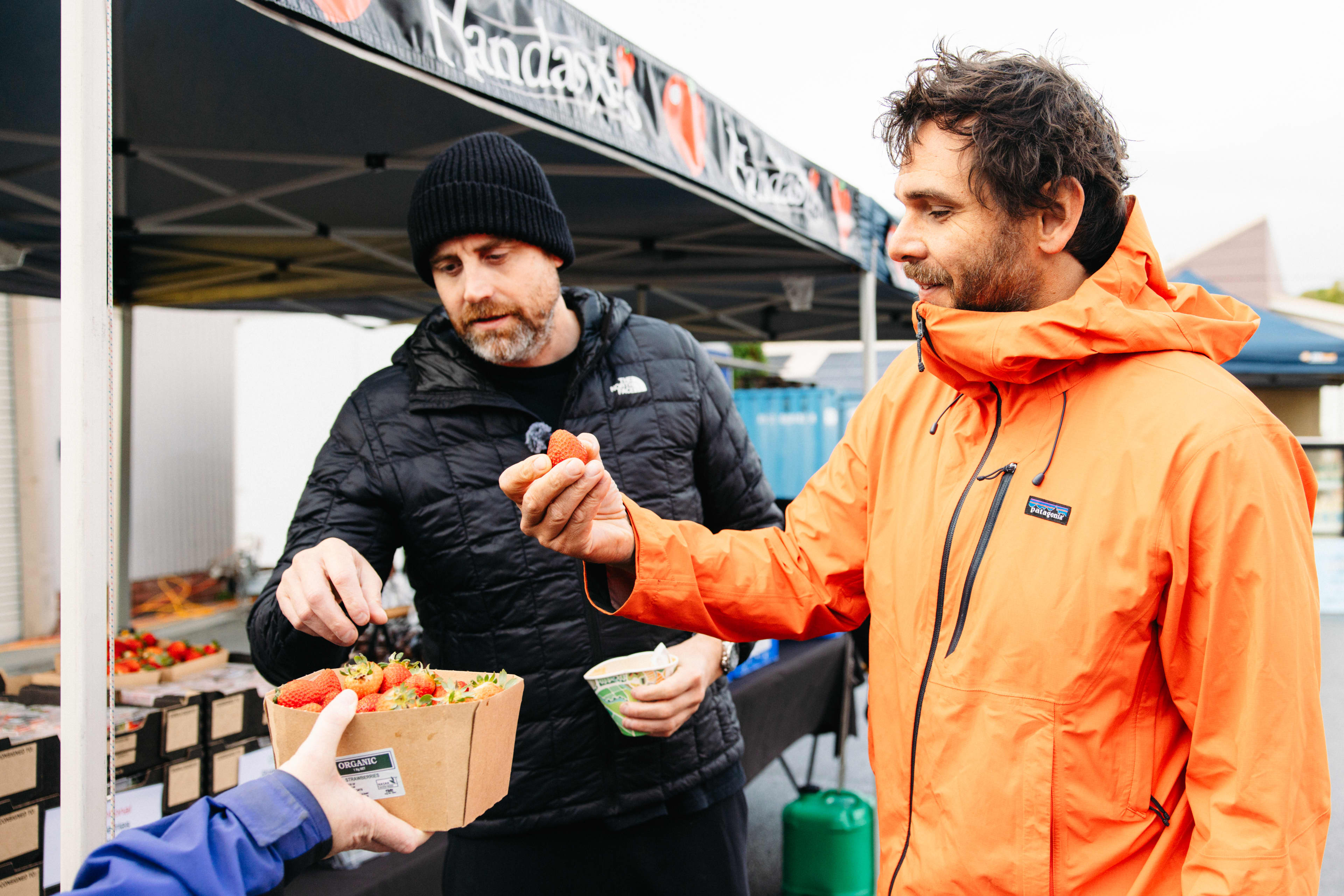 Two people in colourful rain jackets trying food at farmers market
