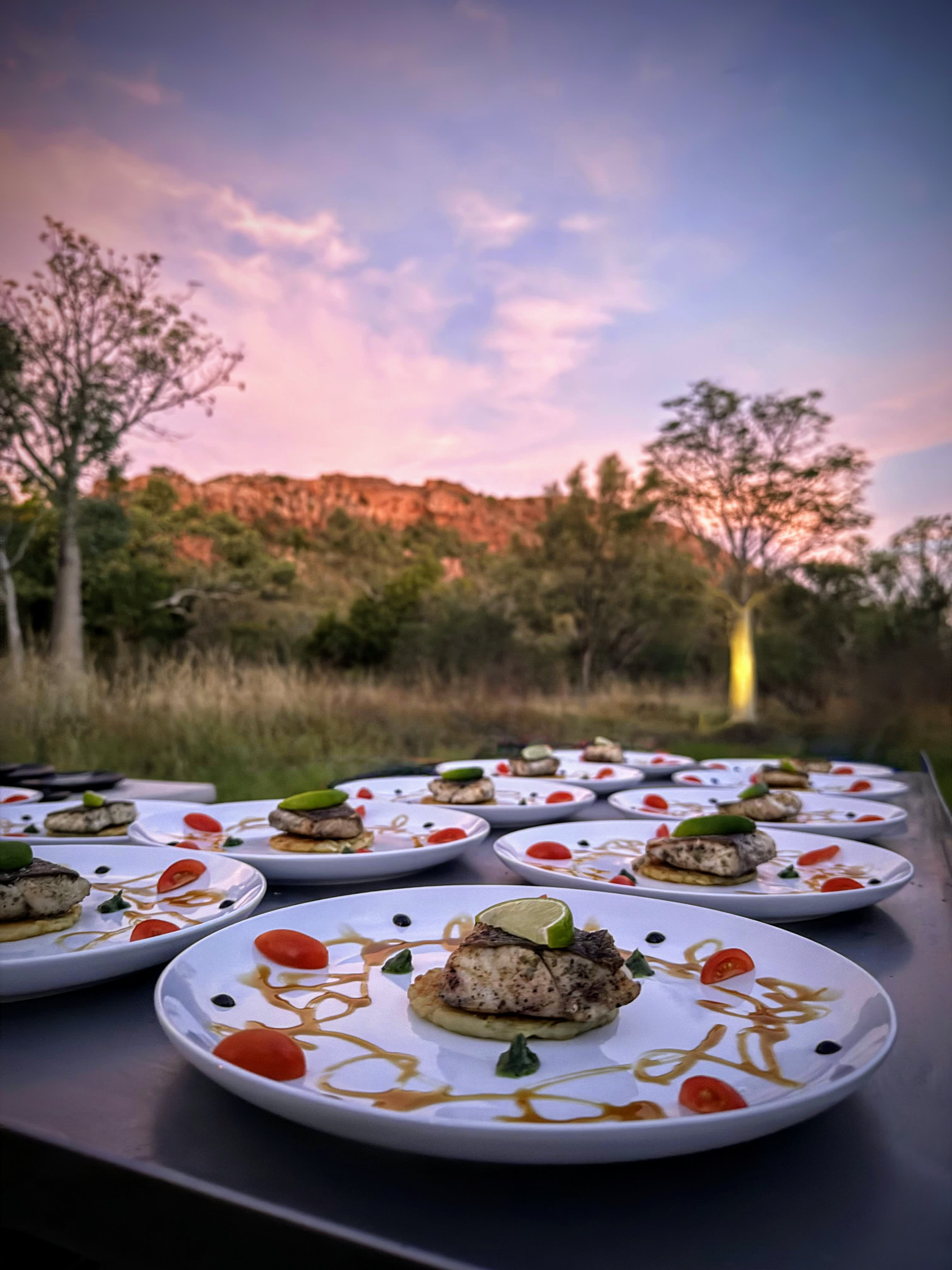 Close up image of food served, with boab trees, red cliffs, and sunset in the background.