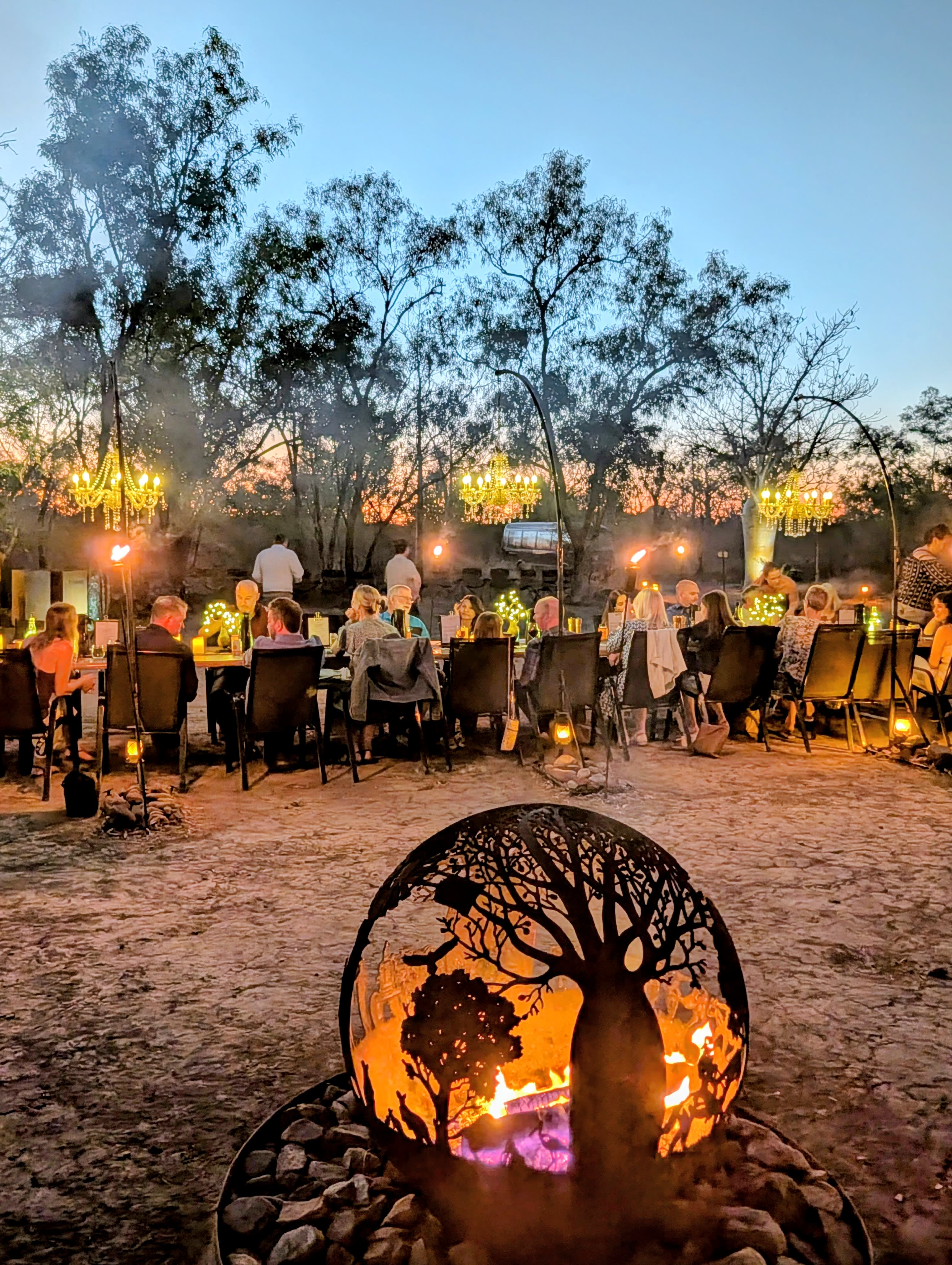 Guests sitting along a long table in the Kimberley outdoors, with a boab open fire pit in the foreground.