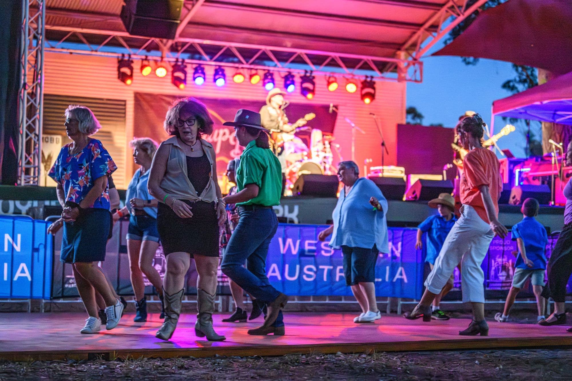 A group of people of various ages dancing in front of an outdoor stage at dusk, with colourful stage lights and a live country band performing in the background at a festival.