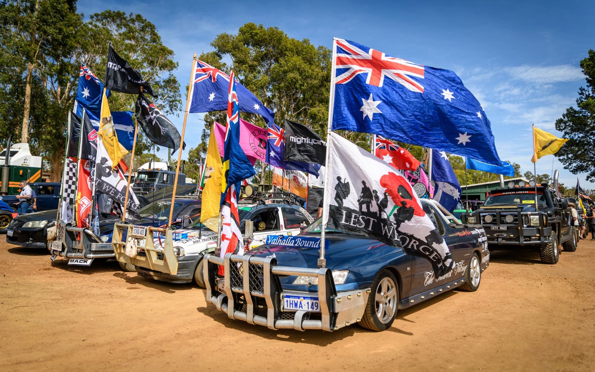 A lineup of rugged bush-bash cars on a dirt road, each decorated with Australian and various other flags.