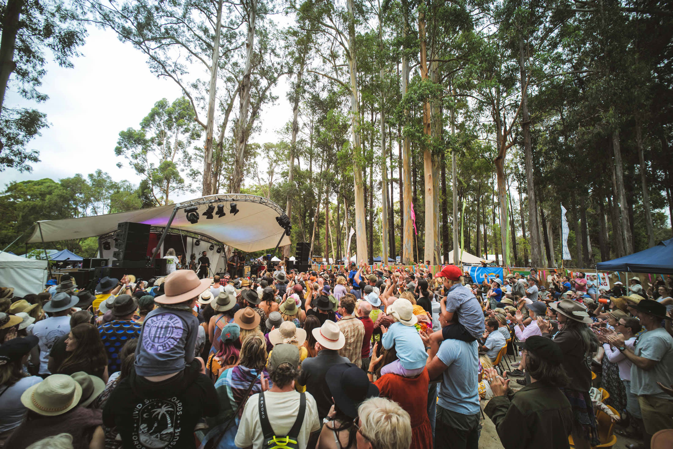 A crowd standing in front of a stage with towering trees surrounding them. A kid with a brown country hat is sitting on the shoulders of his dad.