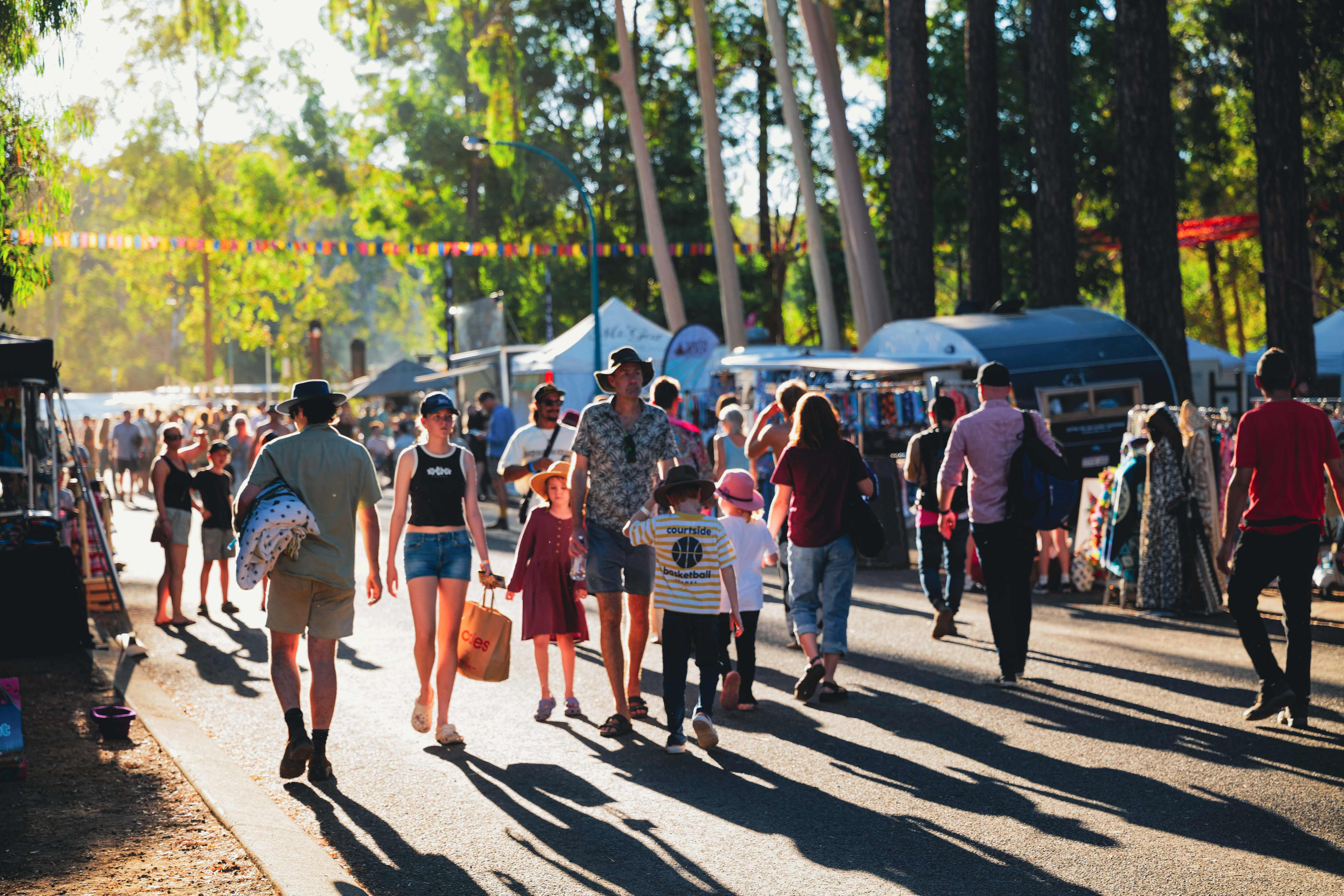 A lively outdoor market scene with many people walking along a sunlit pathway lined with vendor stalls. Adults and children stroll among the booths selling clothing and goods, while tall trees cast long shadows across the ground