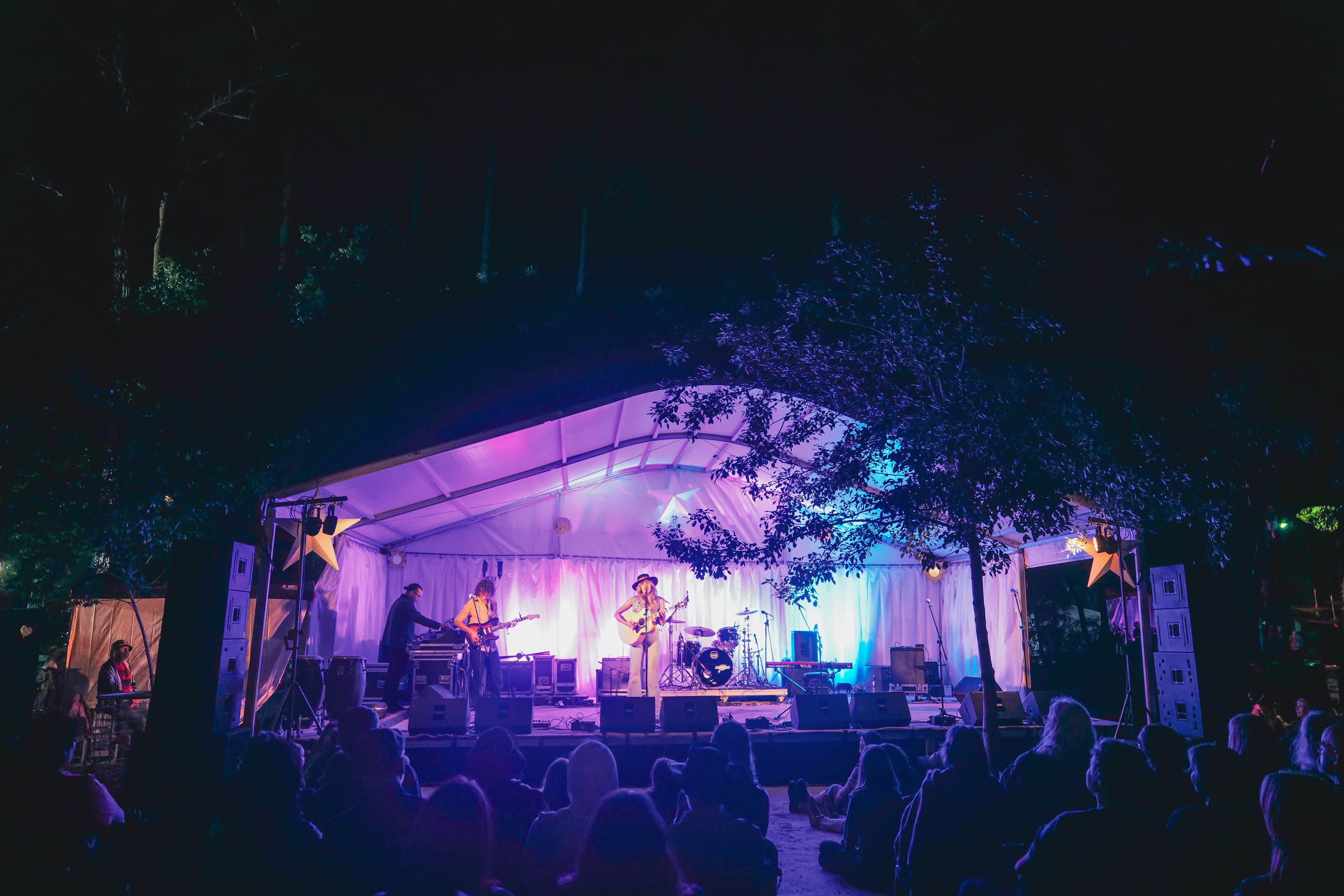 A purple stage with a female standing in front of a microphone with an acoustic guitar, with a crowd sitting in front of the stage