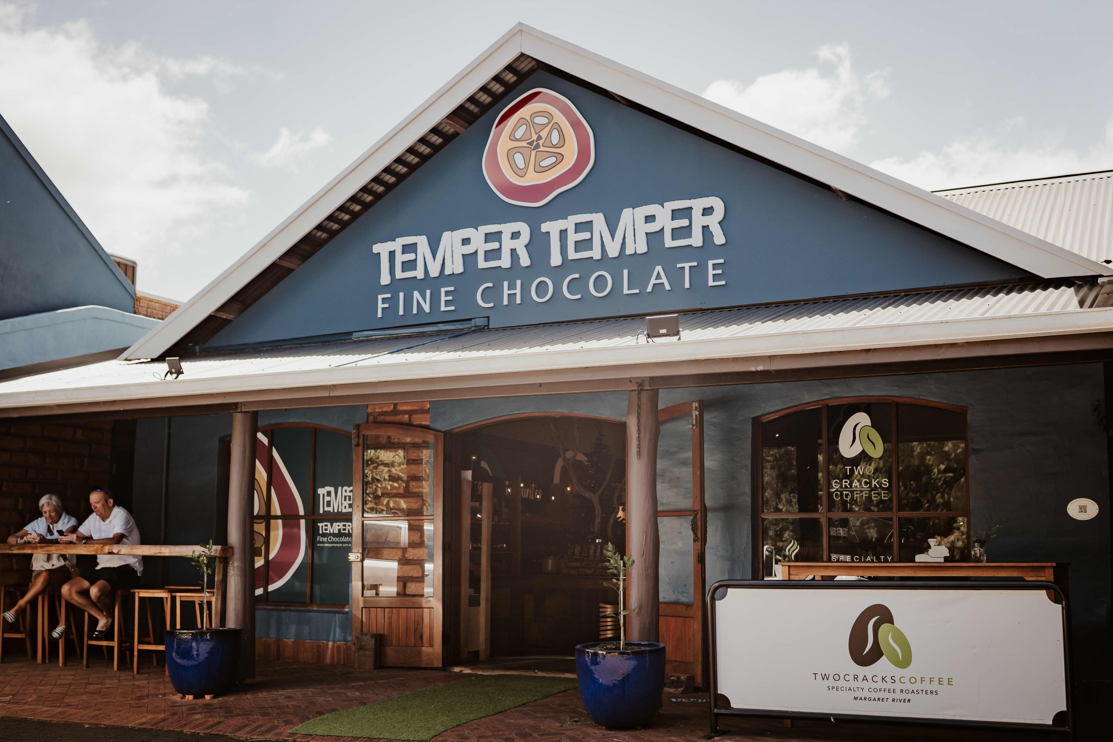 Street view of the Temper Temper Fine Chocolate shop, featuring a blue façade with a large sign and logo above the entrance. To the right is a Two Cracks Coffee counter with its logo displayed on the window and front panel. Two people sit at an outdoor wooden bar on the left, enjoying drinks under the covered patio. The scene is bright and sunny with a relaxed, inviting atmosphere