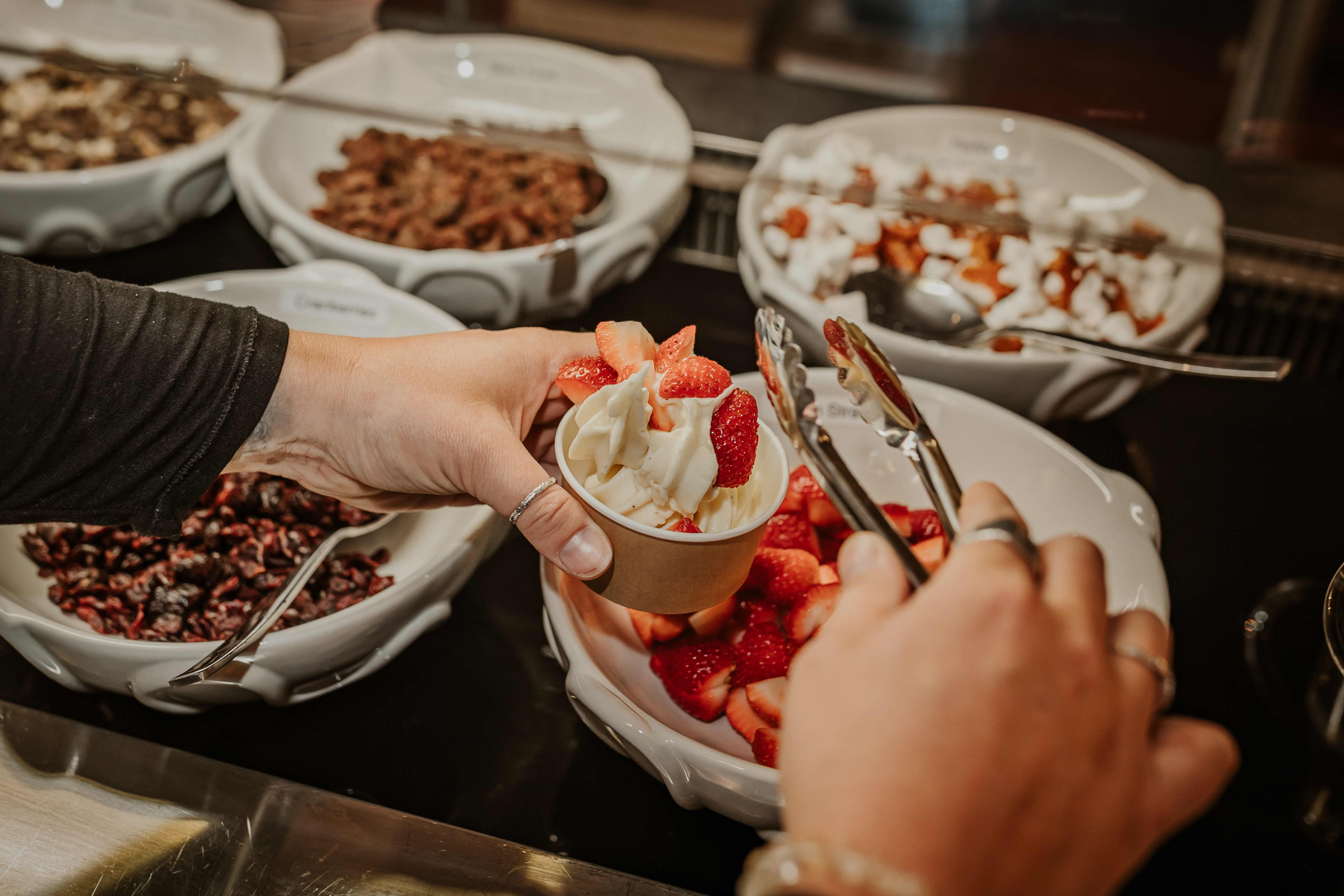 Close-up of a person scooping fresh strawberry pieces with tongs onto a cup of soft-serve ice cream. Several bowls of toppings, including dried fruit and other mix-ins, are arranged on the counter in the background.