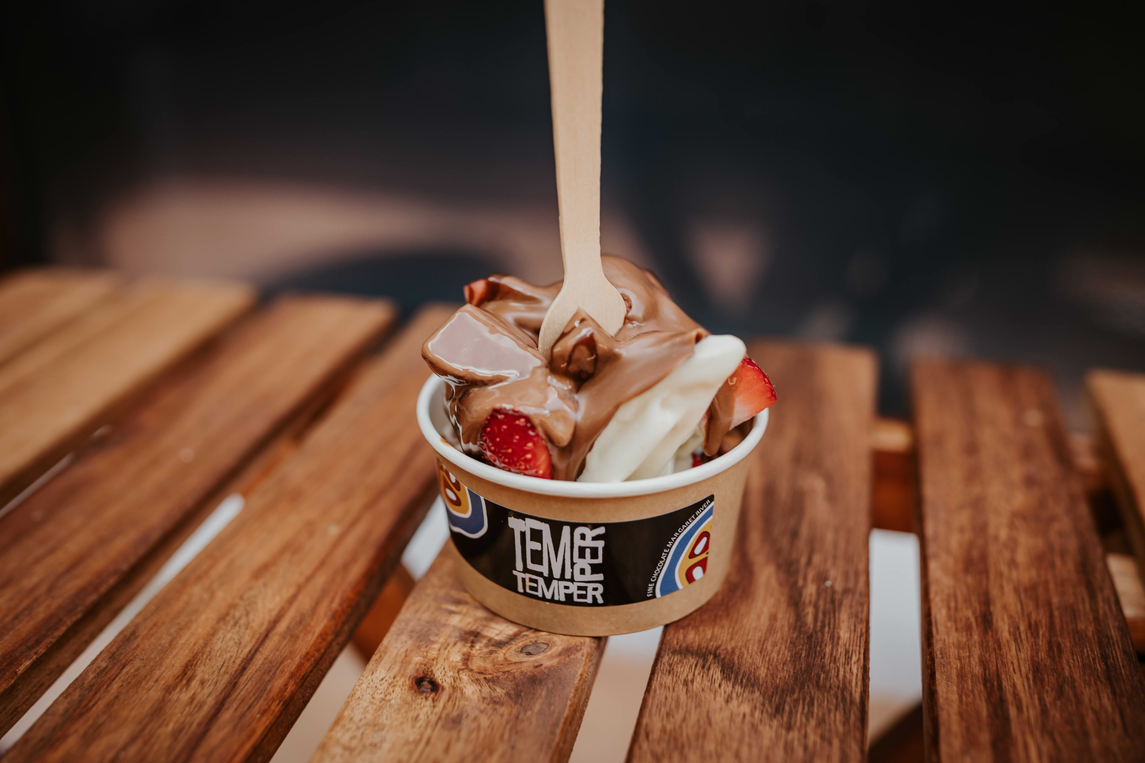 A soft serve ice cream in a cup with melted chocolate and strawberries toppings.