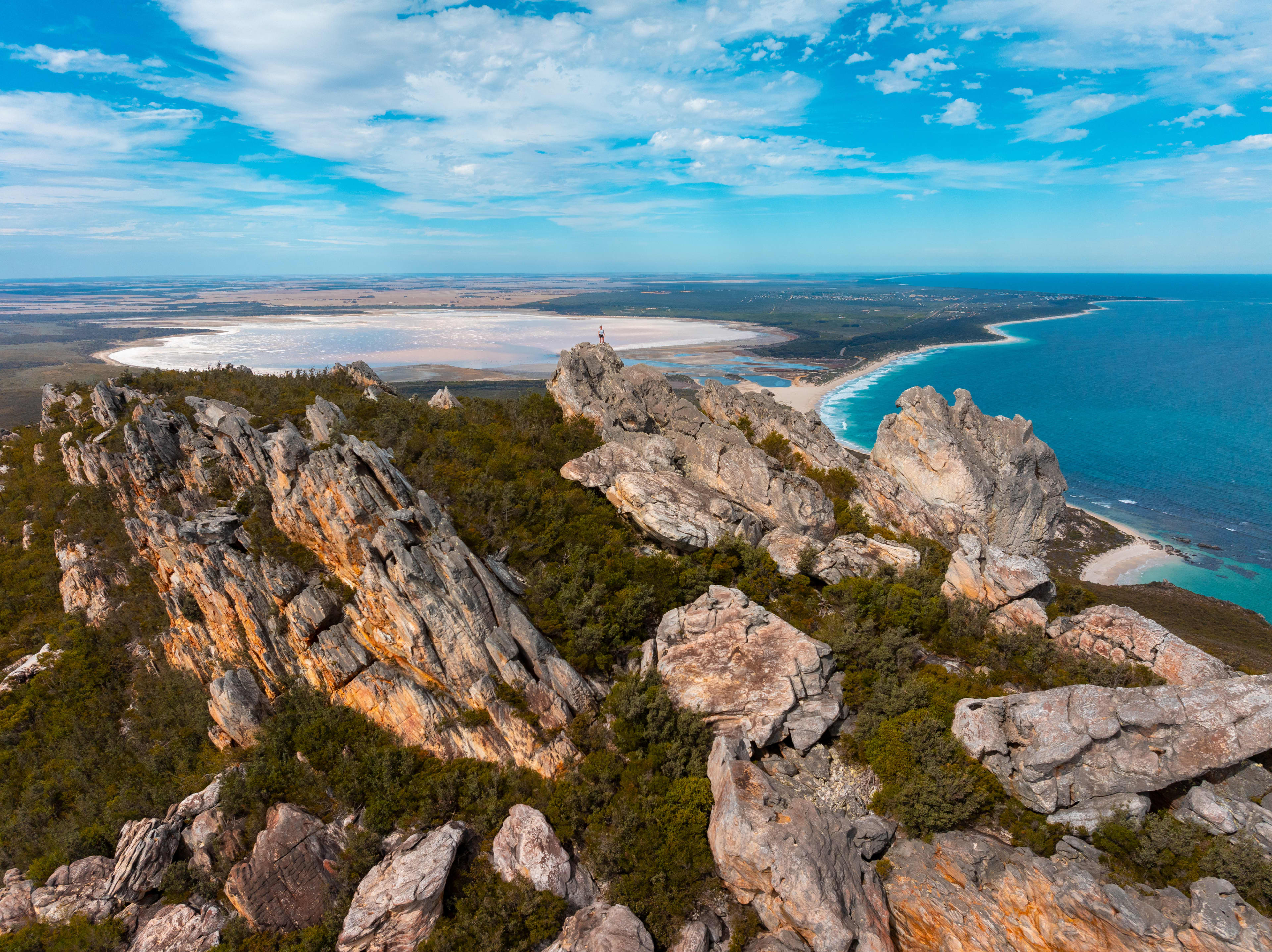 Panoramic view from a rocky coastal summit showing jagged stone outcrops and low green vegetation in the foreground, a sweeping turquoise ocean and sandy shoreline to the right, and a large pale salt lake stretching across the landscape to the left under a bright blue sky with scattered clouds.