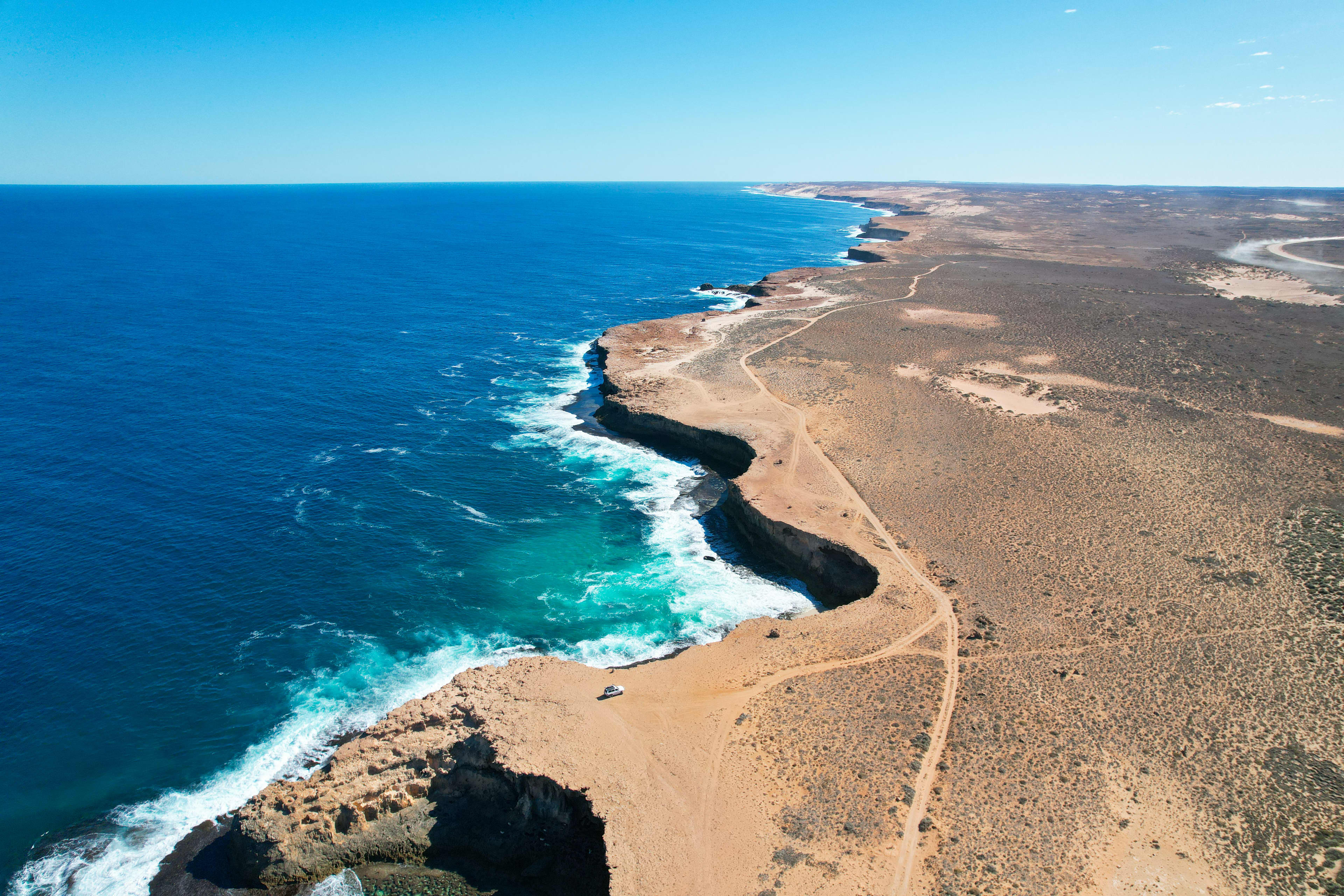 Aerial view of a rugged desert coastline with steep rocky cliffs meeting deep blue ocean waters, white waves crashing below, and sandy tracks winding along the barren shoreline under a clear sky.