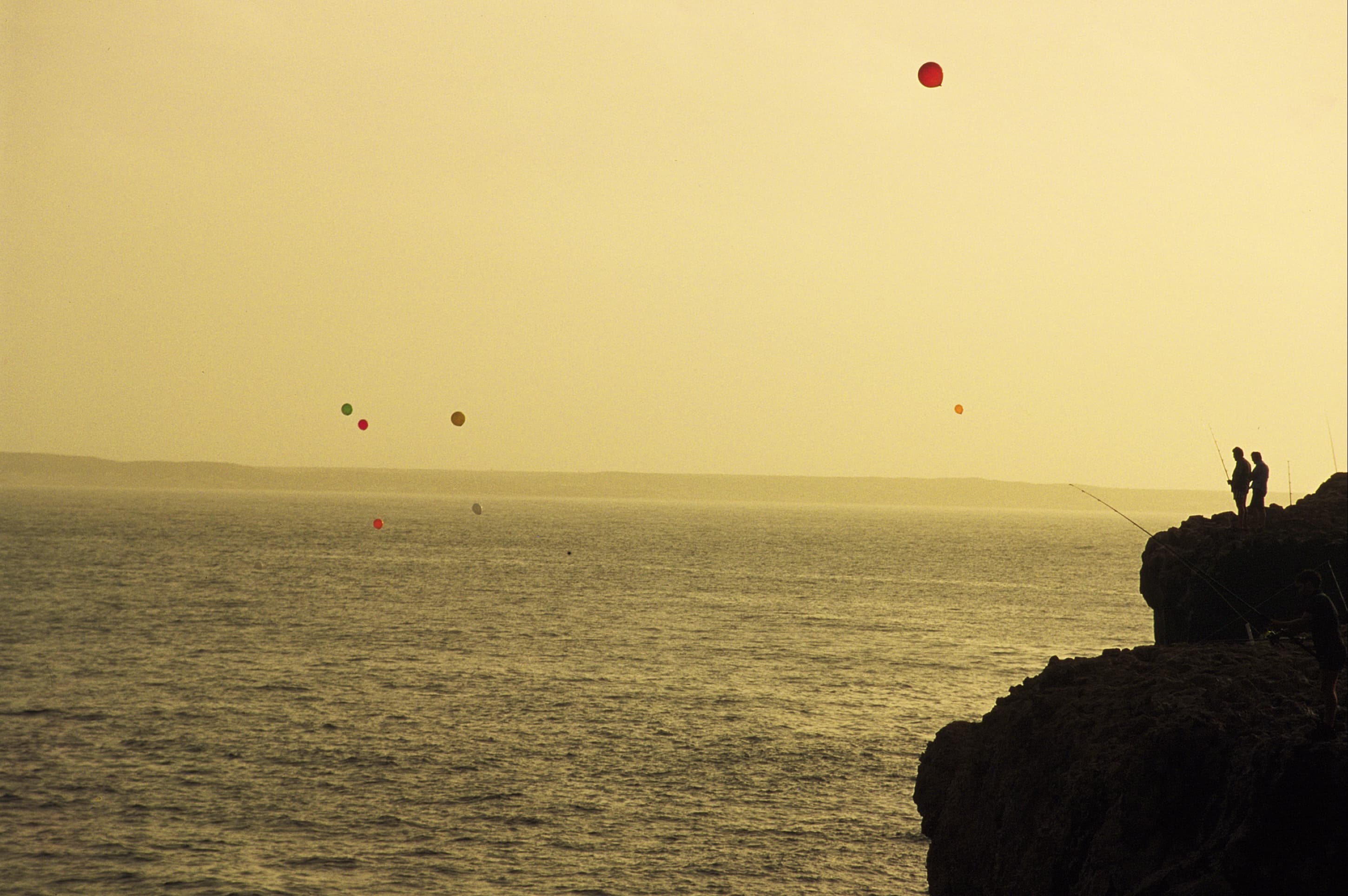 A sunset with two silhouettes of people standing on the edge of a cliff edge fishing with balloons floating in the background