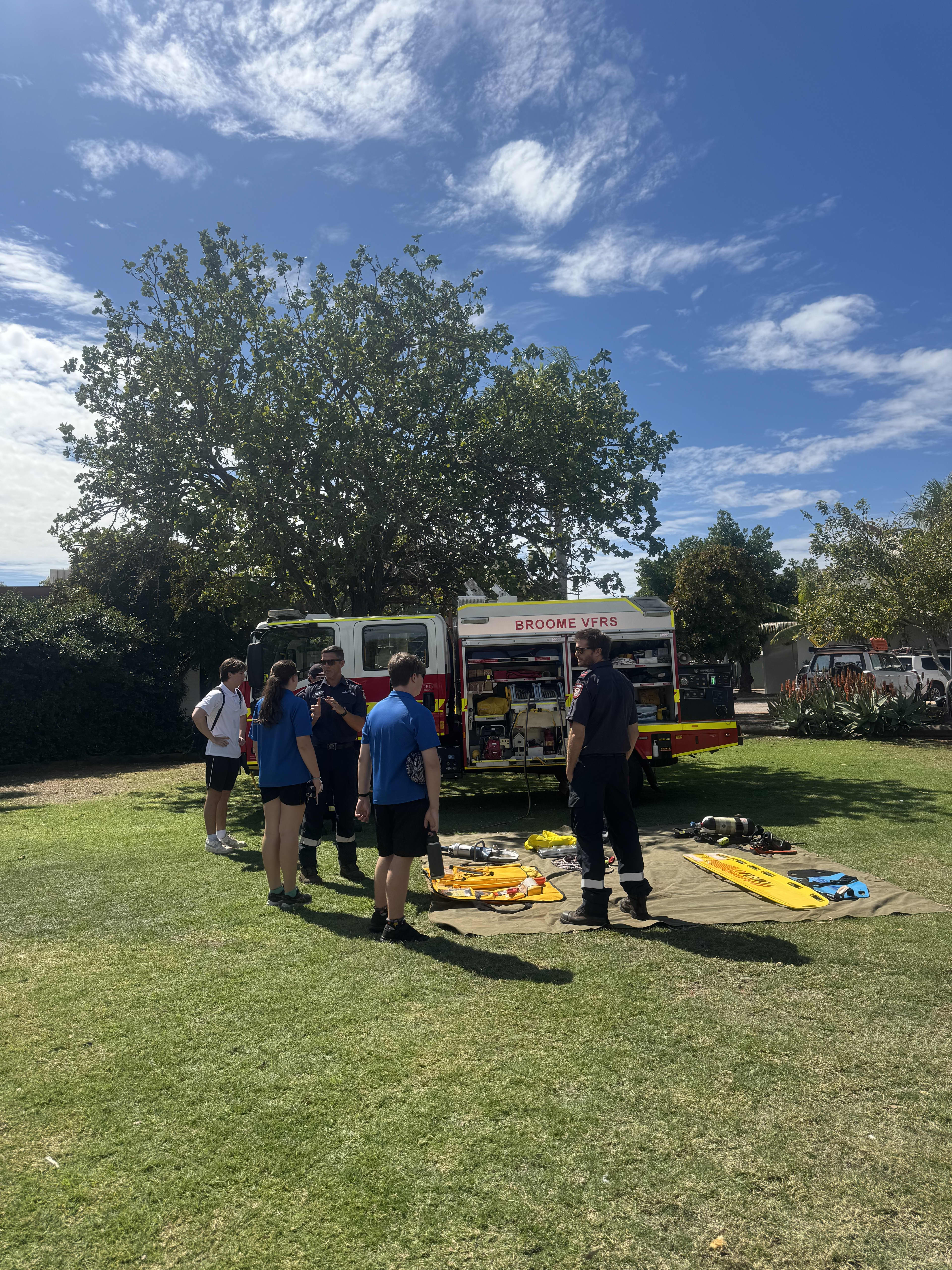 Students standing around Fire truck outside in the green grass and blue sky.